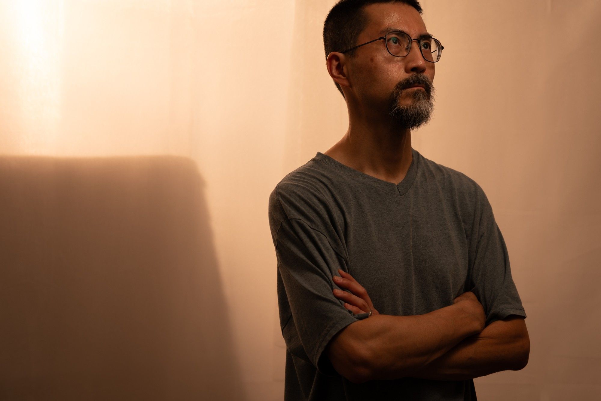 This is a portrait of a middle-aged Japanese man wearing a blue T-shirt. He is looking toward the upper right with his arms crossed. The photograph has an overall orange cast that is suggestive of late afternoon and/or an arid location.