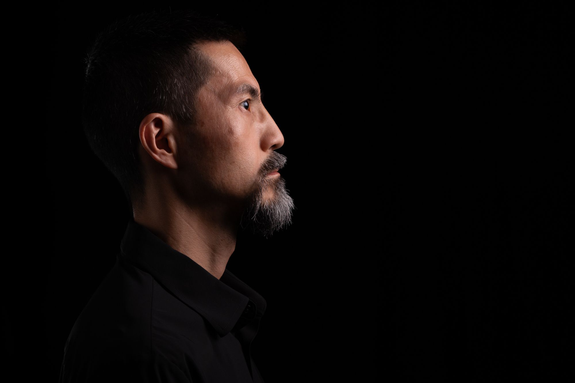 This is a portrait of a middle-aged Japanese man wearing a black shirt in profile view. He is looking to the right. The background is black.