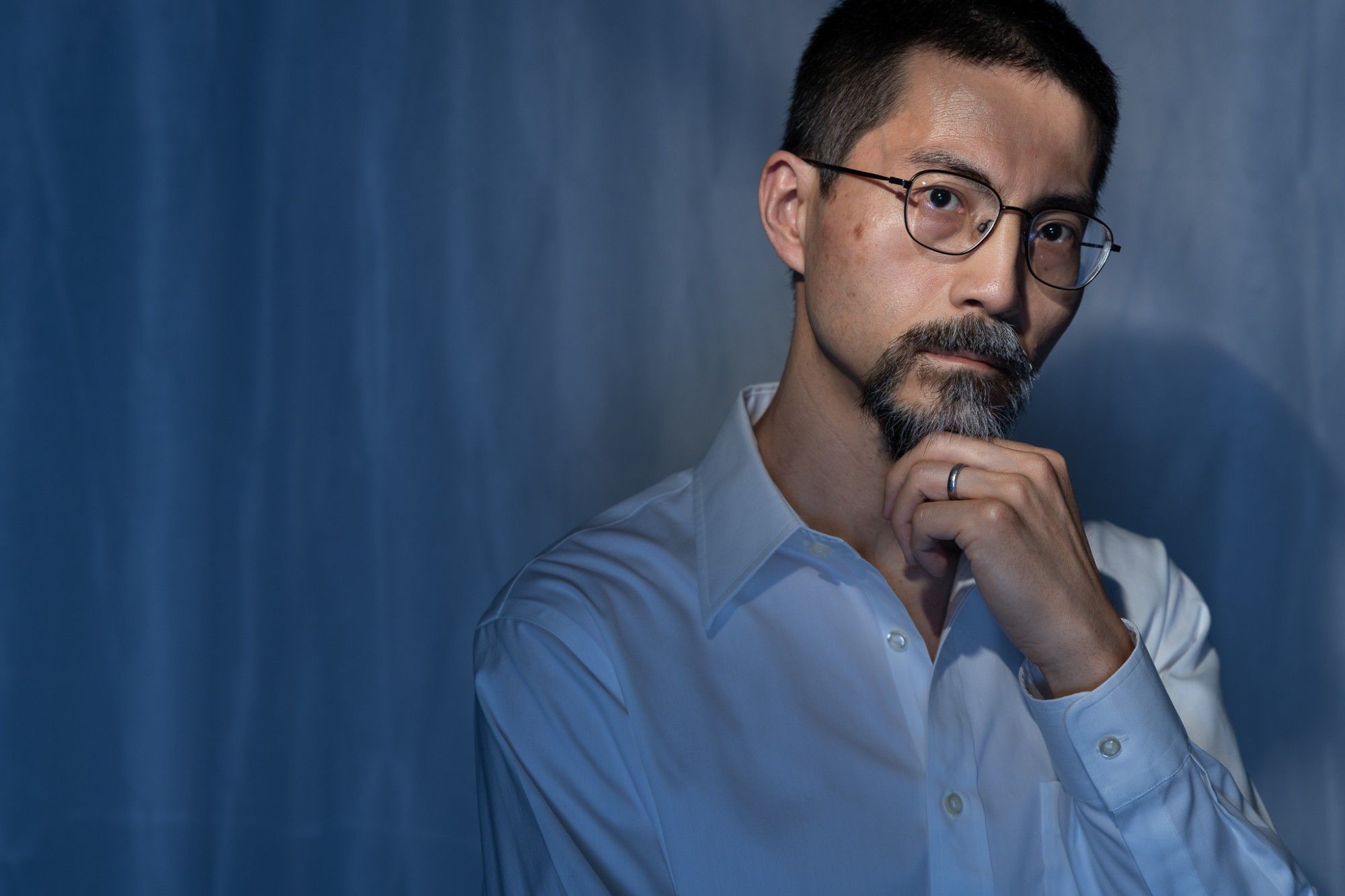 This is a portrait of a middle-aged Japanese man wearing a white shirt. He is wearing glasses and looking straight at the camera while his head is turned toward the right. The background is white but with a blue cast.