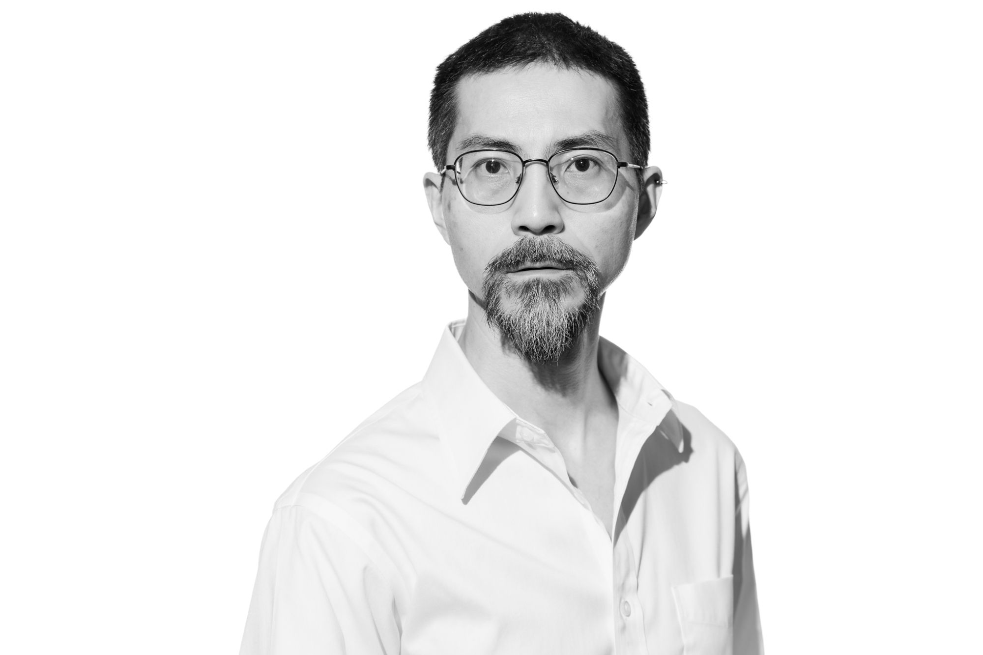 This is a high-key portrait of a middle-aged Japanese man wearing a white shirt. He is wearing glasses and looking straight at the camera. The background is white.