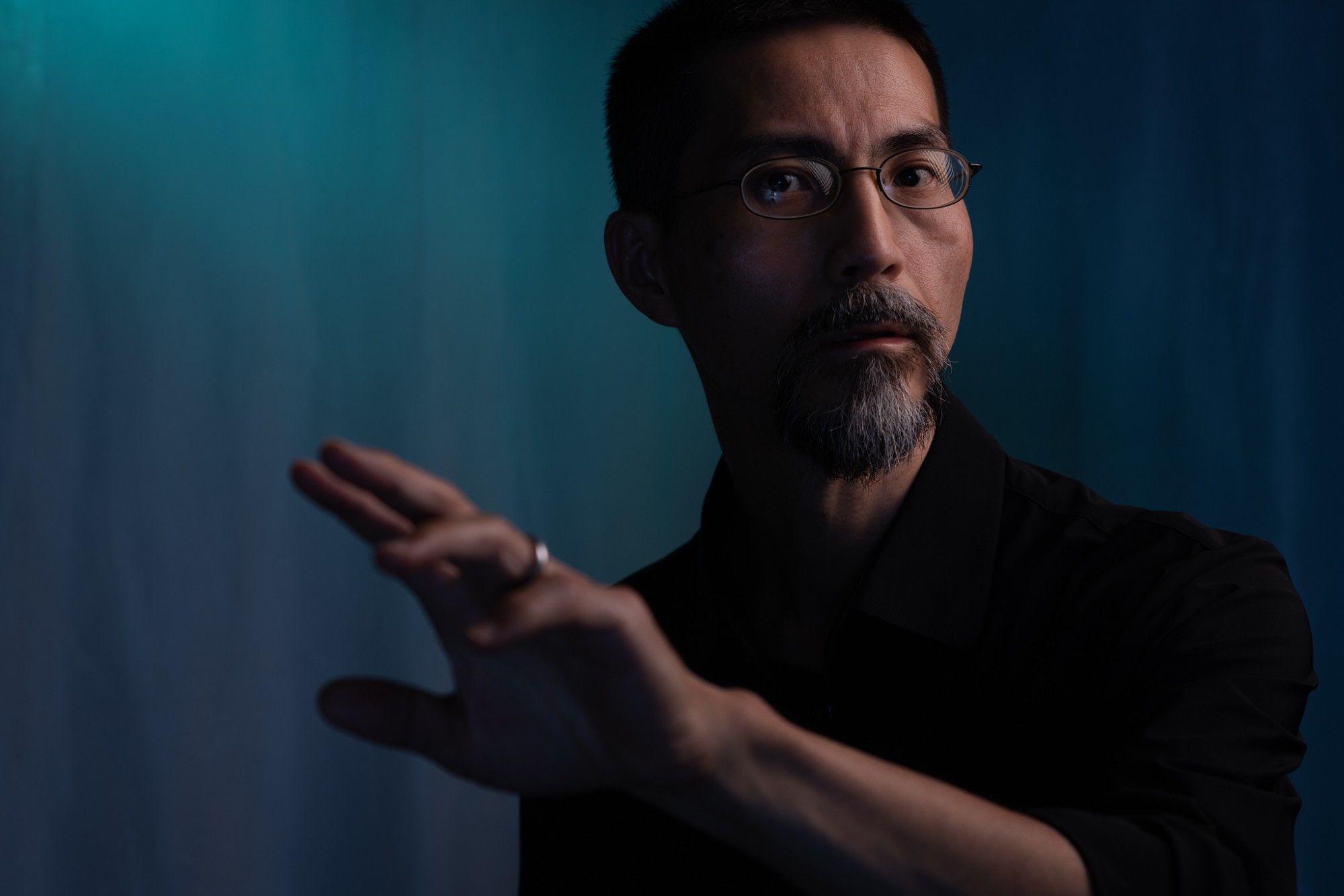 This is a portrait of a middle-aged Japanese man wearing a black shirt. He is looking straight, his head slightly turned toward the right, and his left arm is reaching forward. The background has a blue-cyan cast.