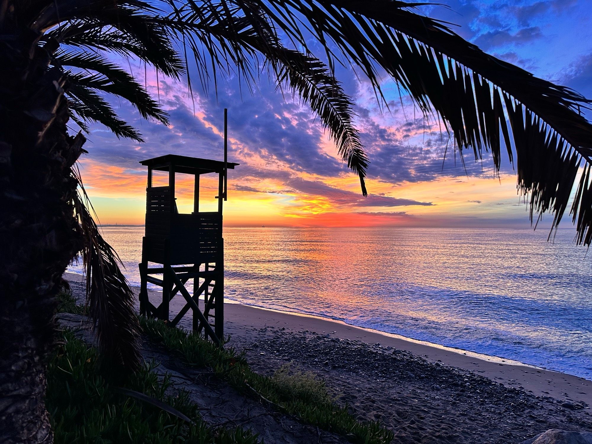 Sonnenaufgang nach seniler Bettflucht in Taragonna, Spanien. Man sieht ein (spanisches) DLRG Wachhäuschen am Strand im Gegenlicht des Sonnenaufgangs, welcher Hinmel und Meer von Miami Platja in allen Farben des Regenbogens leuchten lässt. Im Vordergrund hängt eine dunkle Palme ins Bild 