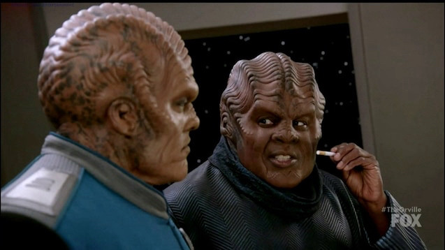 Screencap of The Orville TV show where either Bortus or Klyden (I don’t know which) is holding a cigarette and talking to the other about it, soon to order “500 cigarettes” from the replicator