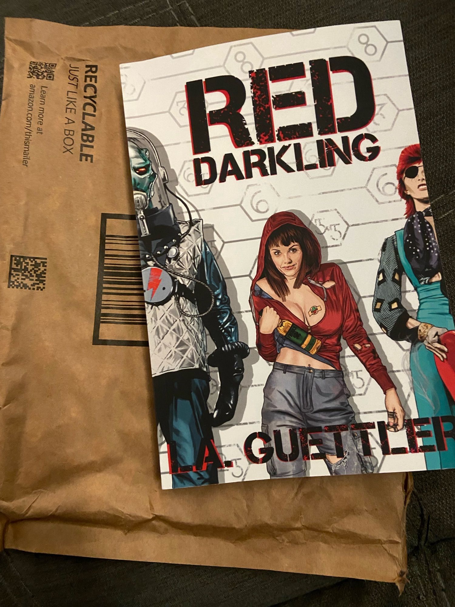 The book Red Darkling by LA Guettler in top of a brown mailing envelope. The cover depicts a woman in a torn, cleavage-baring hoodie snd gray pants holding a bottle of brown liquor, standing in a sci-fi line-up between a tall alien in a crinkly spacesuit and what looks like David Bowie with an eyepatch