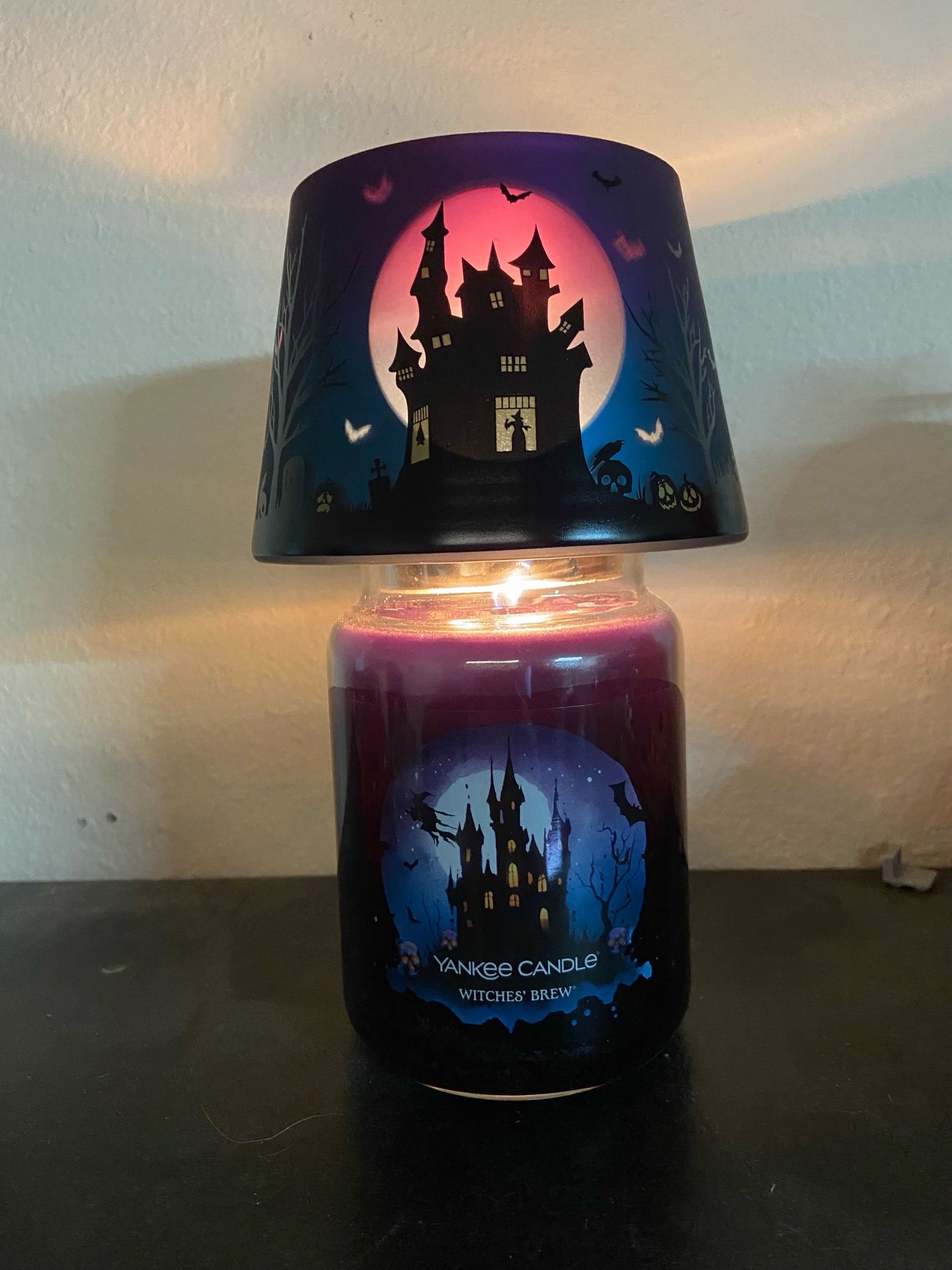 A purple scented candle in a glass jar with a witchy house decor and matching glass lampshade topper, throwing eerie light on the wall