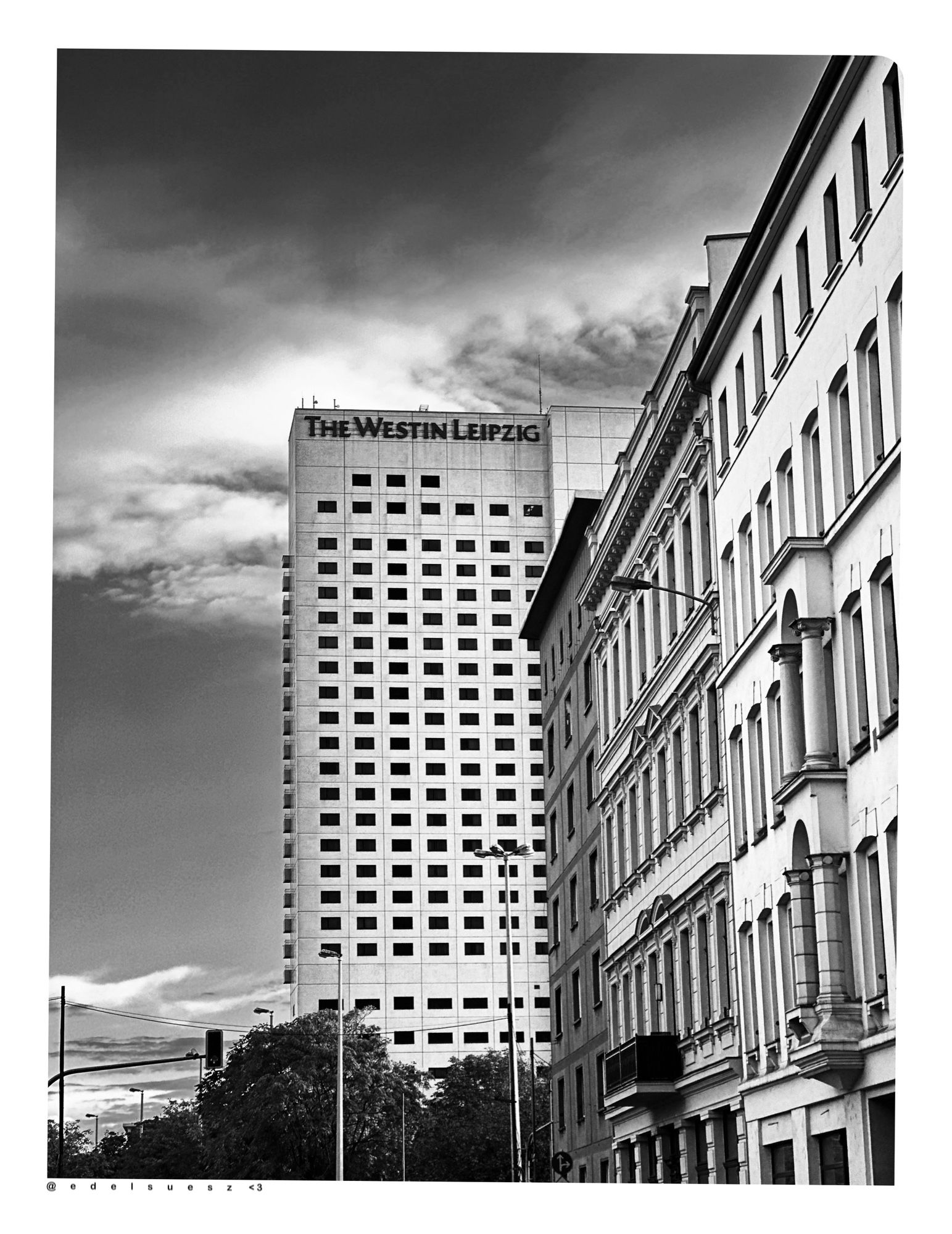 Schwarzweißfotografie: rechts ein alter Straßenzug aus verschiedenen Gründerzeithäusern, dahinter schaut ein Teil des großen, eckigen Hochhauses heraus, das ehemalige Hotel Merkur, jetzt The Westin Leipzig, unzählige kleine, aus der Entfernung schwarze, viereckige Fenster, helle Fassade plus großer Schiftzug THE WESTIN LEIPZIG, weiße Wolken darüber und grauer Himmel im Hintergrund, im unteren Bereich eine Ampelanlage, Straßenlaternen und mehrere Bäume