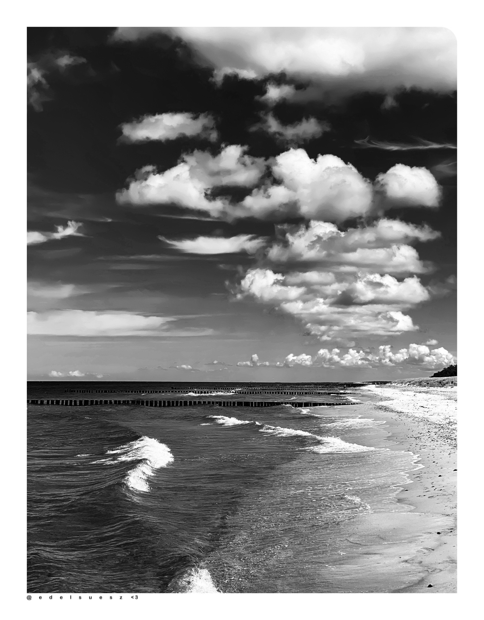 Schwarzweißfotografie: Blick seitlich auf einen langen hellen Sandstrand, links die tosenden, schaumig hellen Wellen, auf den Strand treffend, unterbrochen von Holzbuhnen, Küstenschutzbauwerke, in Reihen gerammte Holzstämme, über dem Horizont grauer heller Himmel mit kleinen unterschiedlich geformten weißen Wolken