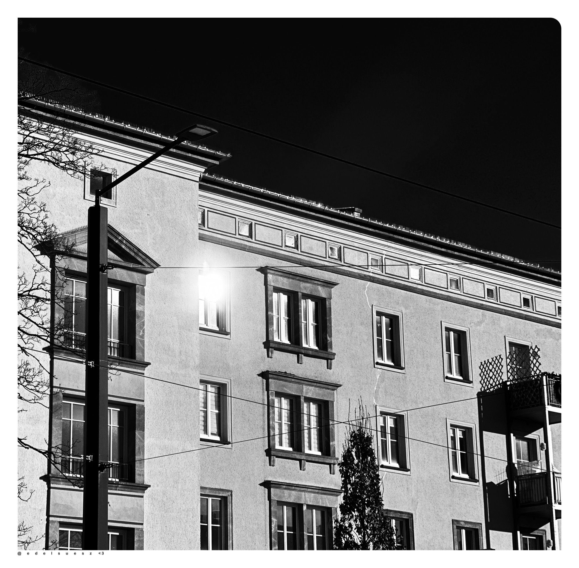 Schwarzweißfotografie: mehrstöckiges, großes Wohngebäude, Stil: Sozialistischer Klassizismus, viele Fenster, in einem spiegelt sich grell das Sonnenlicht und blendet, links eine Straßenlaterne, rechts vorgesetzte Anbaubalkone, darüber einheitlicher dunkler Himmel