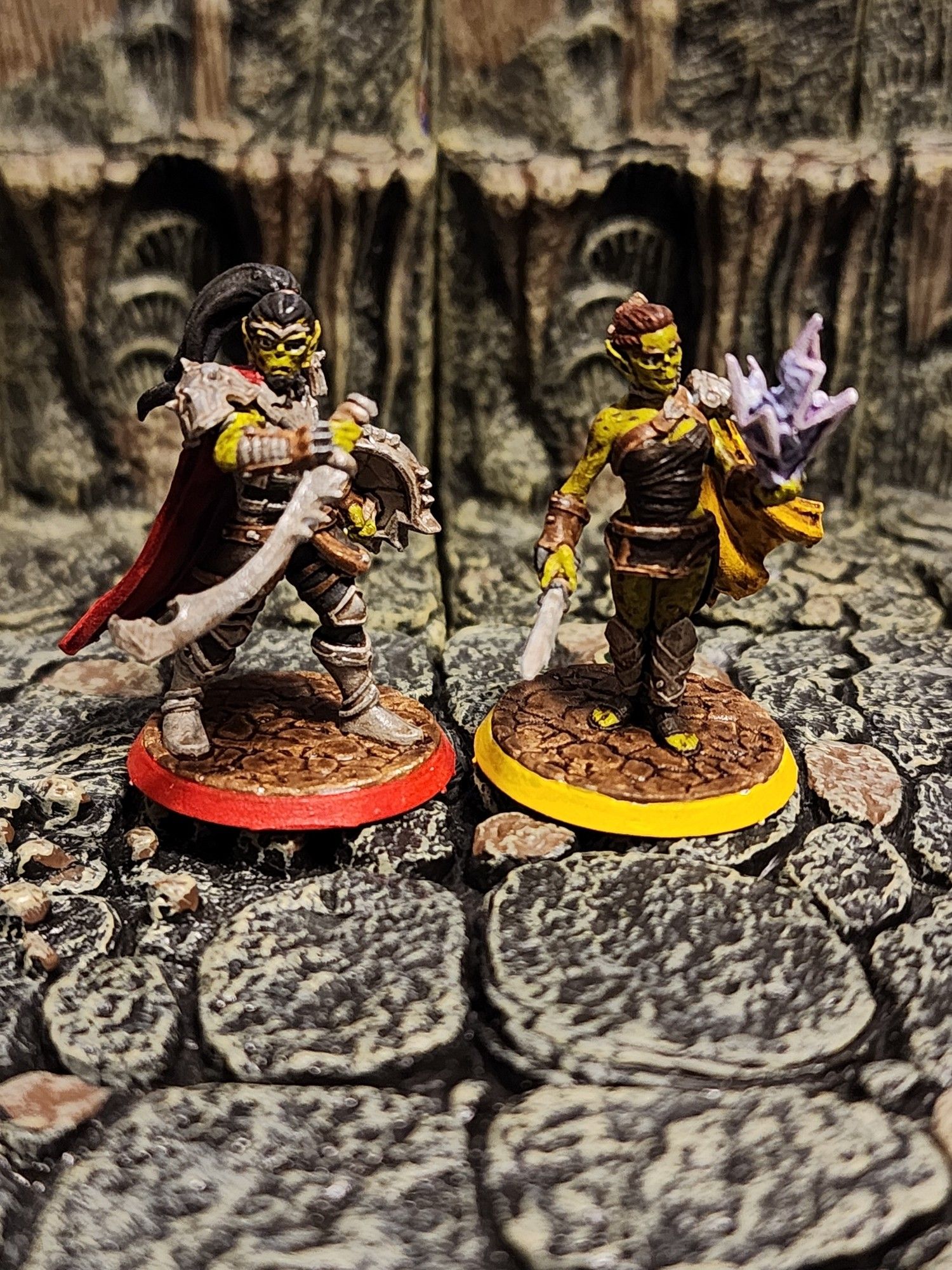 Two custom designed and painted Githyanki miniatures. The one on the left is masculine, with long black hair, silver armor, a curved sword, a spiked shield, and a red cape. The one on the right is feminine, with short auburn hair, brown leather armor, a long dagger, a yellow half-cape, and a burst of magical lightning dancing at her fingertips.