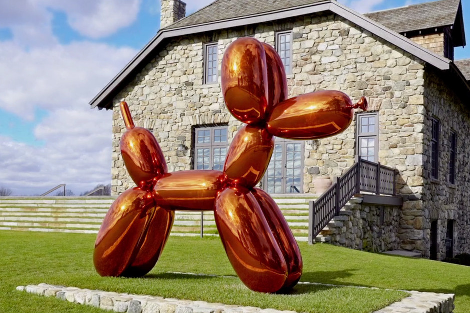 Large reddish orange balloon dog statue outside on a green grass lawn with a large stone house in the background.
by Jeff Koons