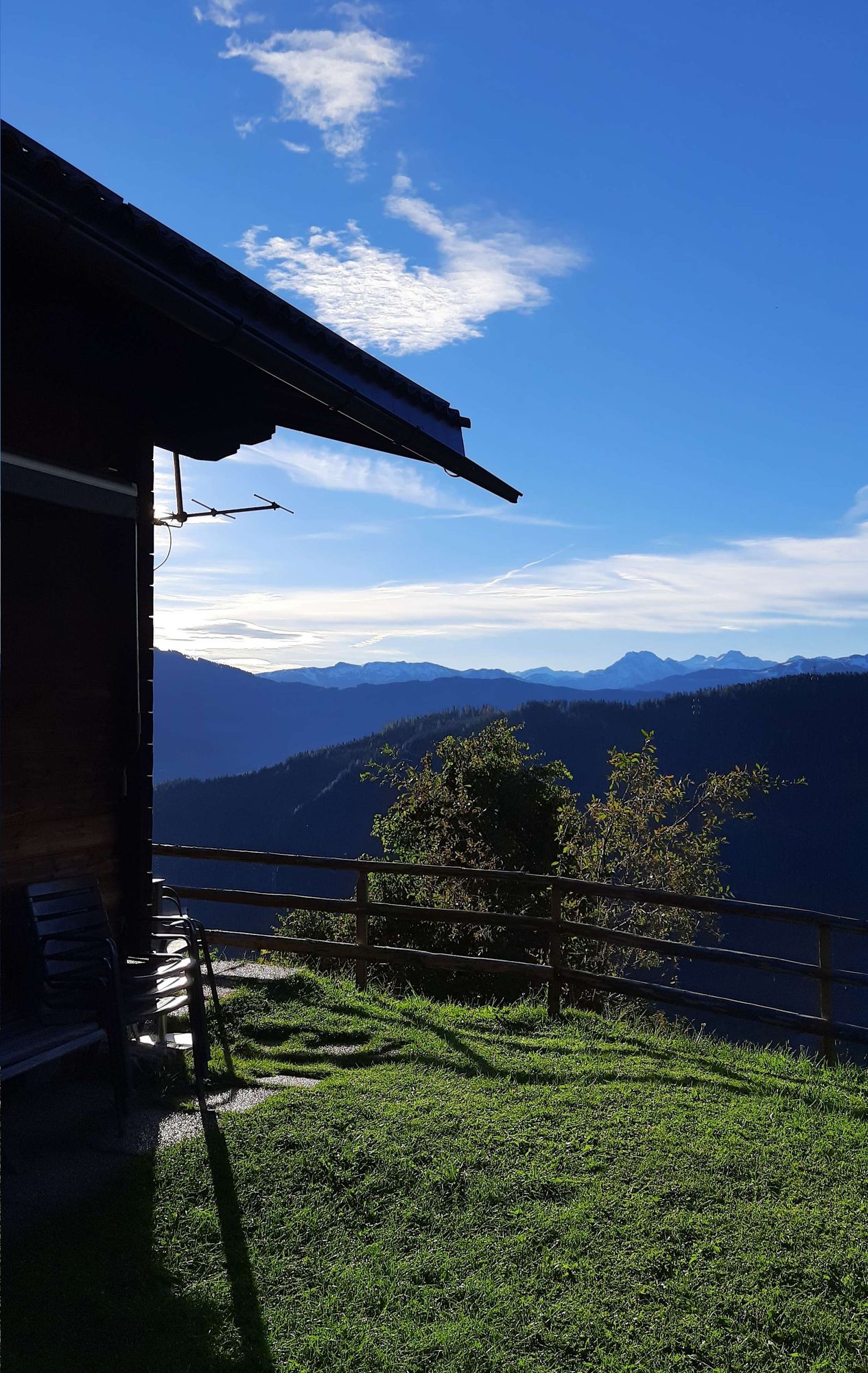 Blick auf die angerissene Silhouette einer rustikalen Berghütte im Gegenlicht der Morgensonne. Rechts Wiesengrün, mittig ein Querlattenzaun, dahinter Weitblick ins Bergpanorama. Ein paar versprengte Schleierwolken kreuzen das Himmelblau.