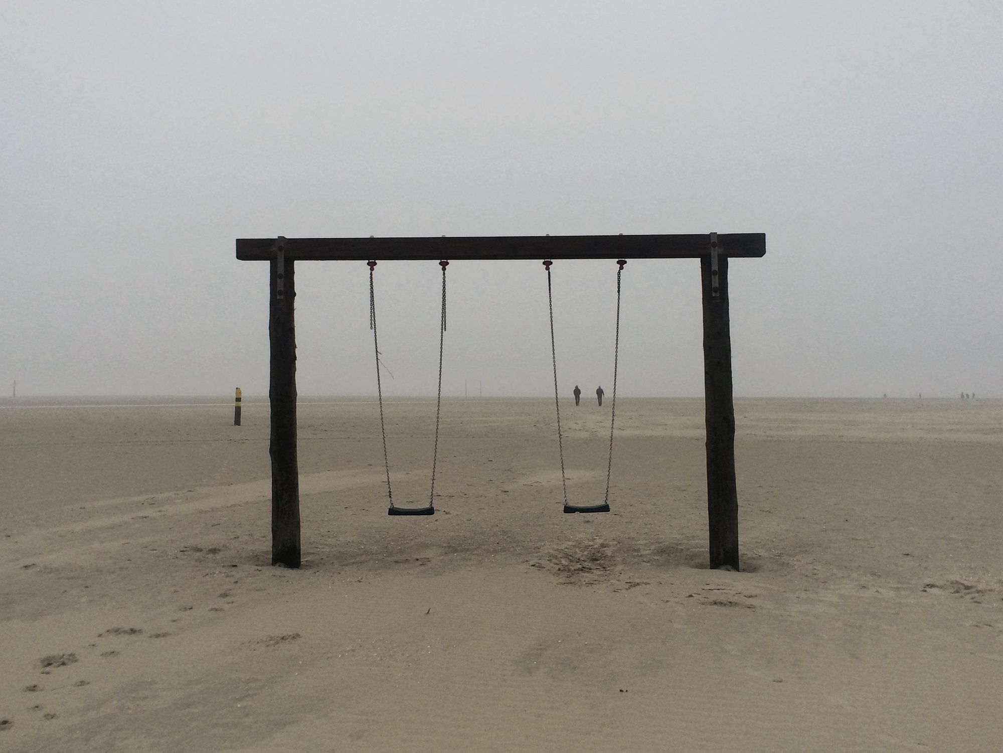 Am neblig-diesigen Strand von St. Peter-Ording steht ein Schaukelgerüst aus dunklem Holz und zwei Schaukeln, die an Ketten hängen. In der Ferne sind zwei Menschen zu sehen, die sich genau zwischen den Ketten der rechten Schaukeln befinden. 