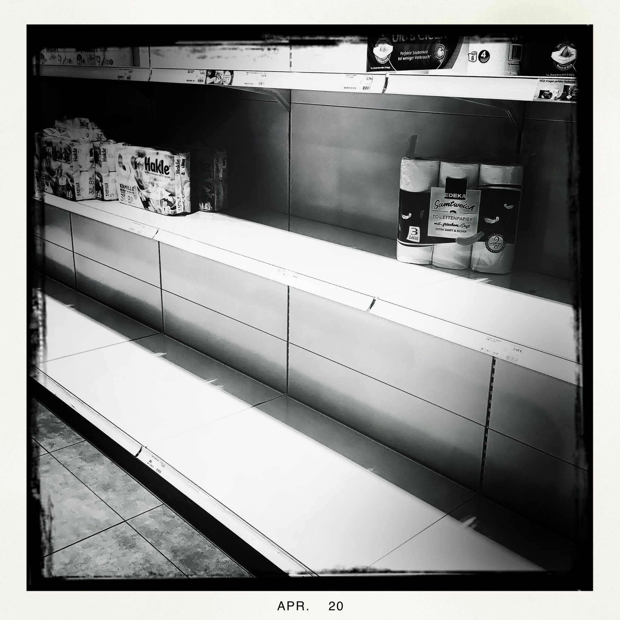 S/W-Bild von fast leeren Supermarkt-Regalen, in denen nur noch ganz vereinzelt wenige Packungen Toilettenpapier stehen. Entwickelt mit Hipstamatic (John S/BlacKeys BW).