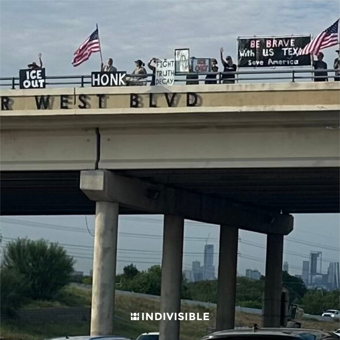 A group of demonstrators displaying various signs and American flags on an overpass above a highway, with a city skyline in the background. Signs include phrases like "Fight for Truth," "Be Brave Be with us," and "Save America." The word "INDIVISIBLE" is visible at the bottom of the image.
