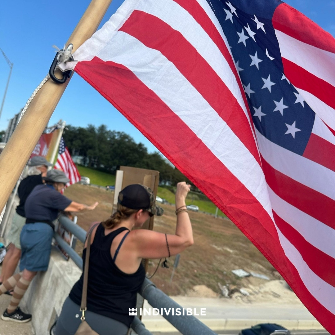 Person waving an American flag over a bridge railing with other flags and people in the background, under a clear blue sky.