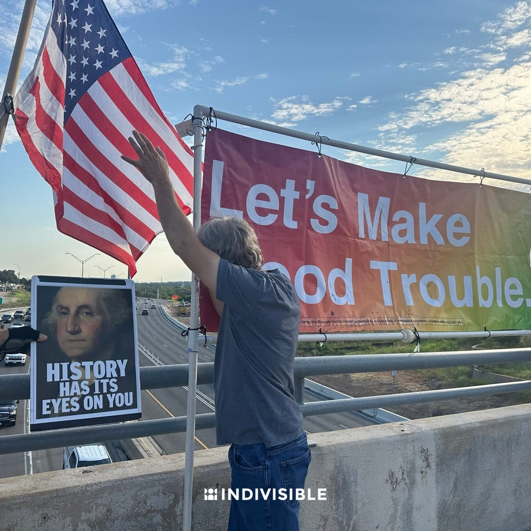 Person waving to a large American flag and a banner reading "Let's Make Good Trouble" on a bridge railing, with a portrait of George Washington and the logo of Indivisible.