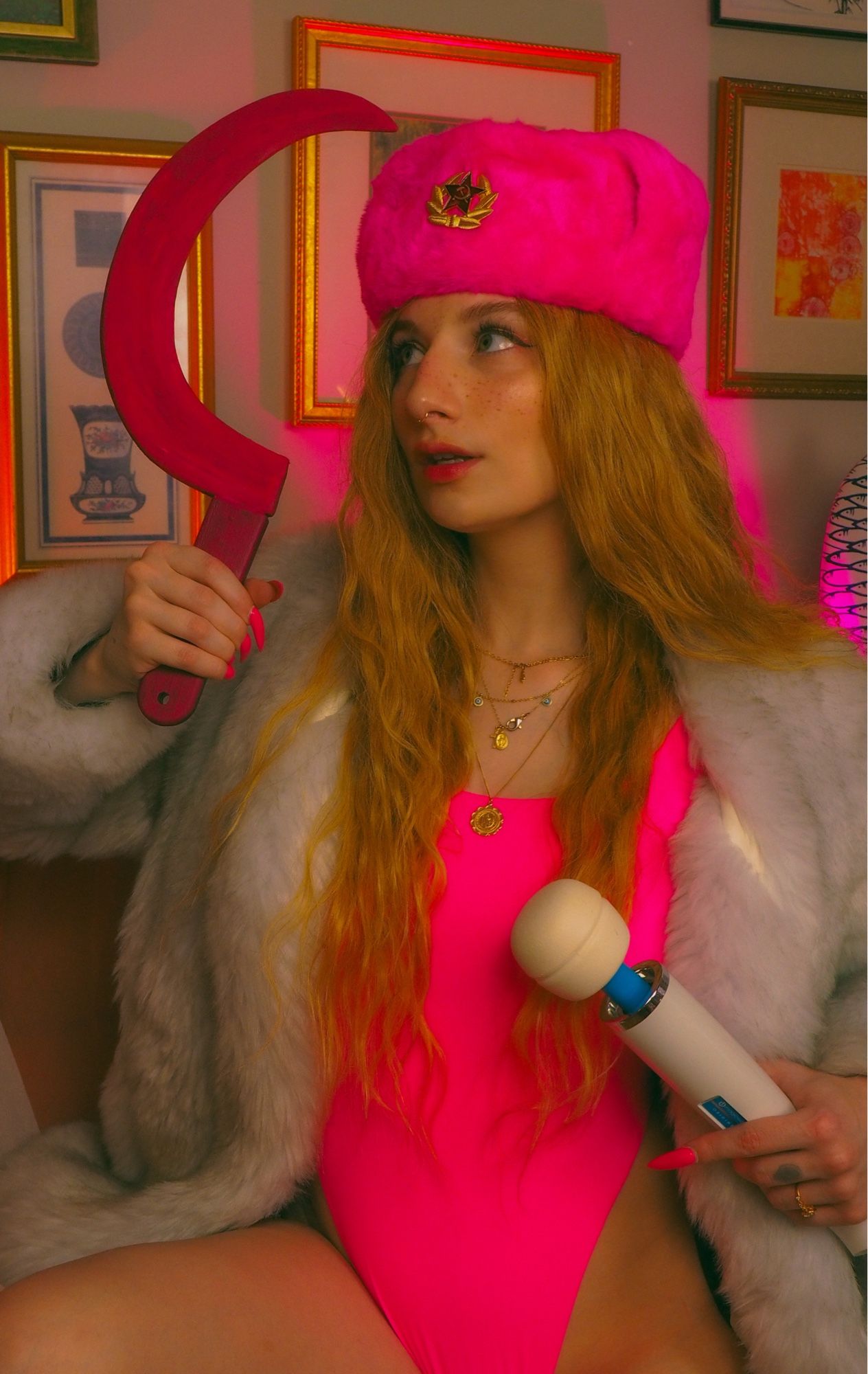 marina in a hot pink ushanka and matching body suit. also wearing a white fur coat. they’re dramatically posed holding a pink sickle and hitachi magic wand