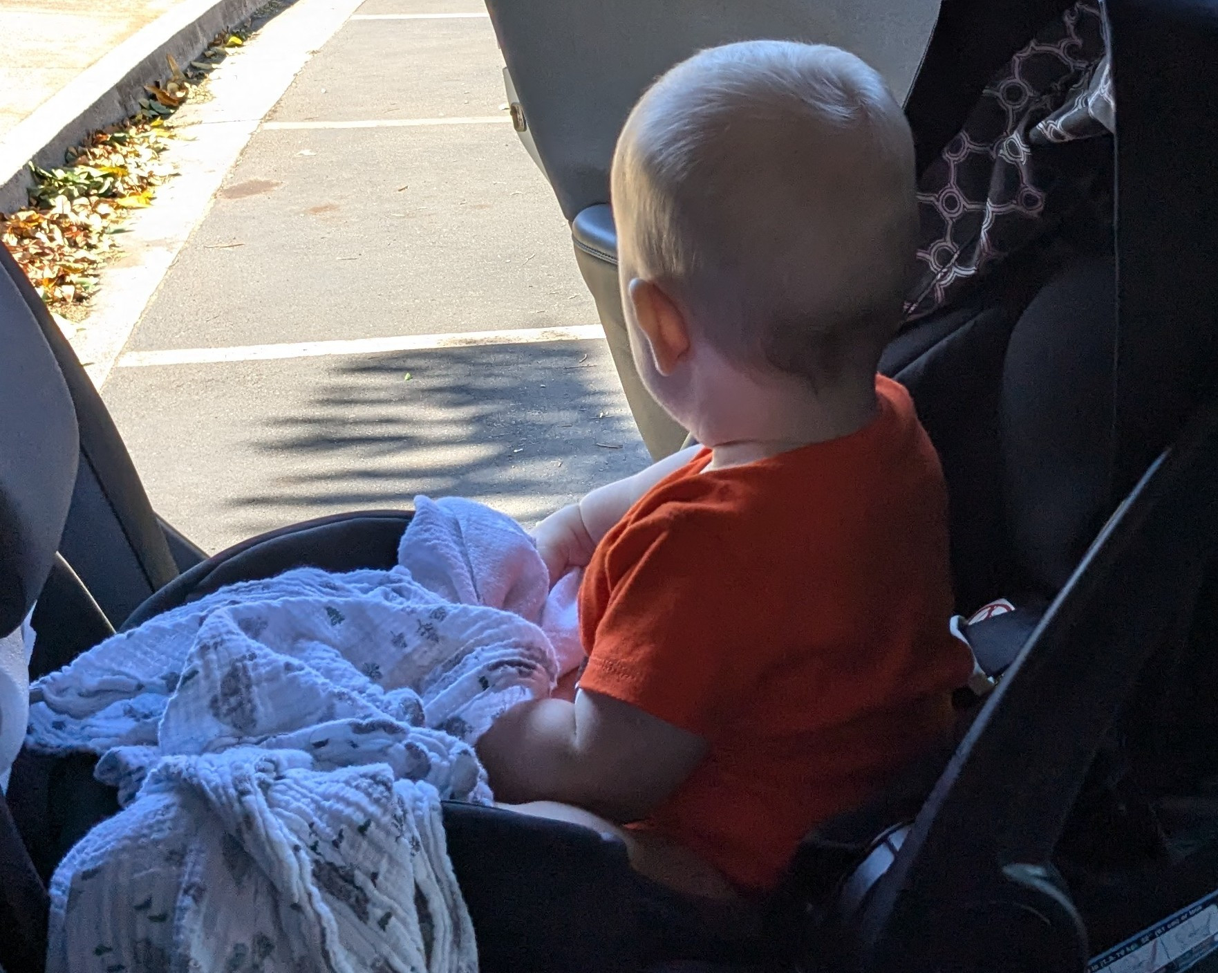 Baby in orange shirt sitting straight up in a car seat. The car door is open and they are looking out