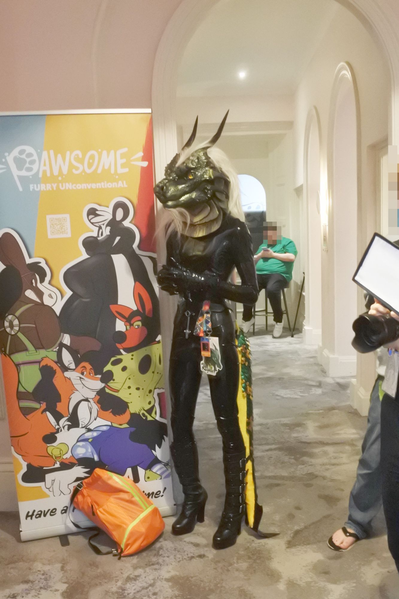 Fursuit photo - a green dragon in a shiny rubber catsuit with claw gloves and leather boots watches and waits as another fursuiter is photographed on the hotel stairs (to the left, off-shot)