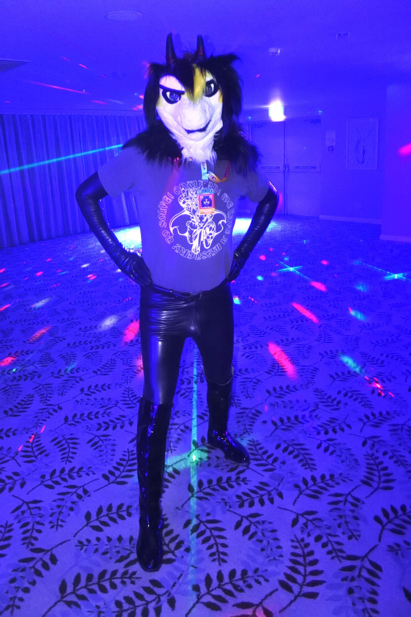 Fursuit photo of a black white and yellow dragon at Pawsome.  He is wearing latex opera gloves, latex leggings and pvc boots.
He's standing on the dance floor, flooded with blue light with a couple of lasers behind him.  This gives the image a blue tint and the latex is shining blue as well.

Photo by Mutley