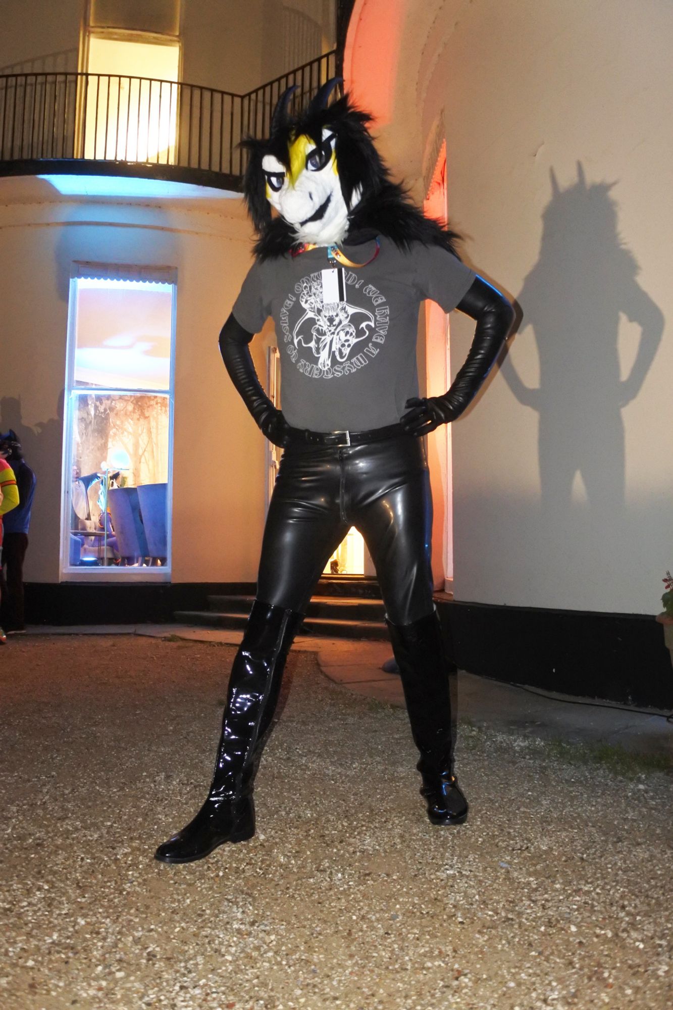 Fursuit photo of a black white and yellow dragon at Pawsome.  He is wearing latex opera gloves, latex leggings and pvc boots.  He's standing outside at night and the outdoor lights are casting his shadow onto the wall.

Photo by Mutley