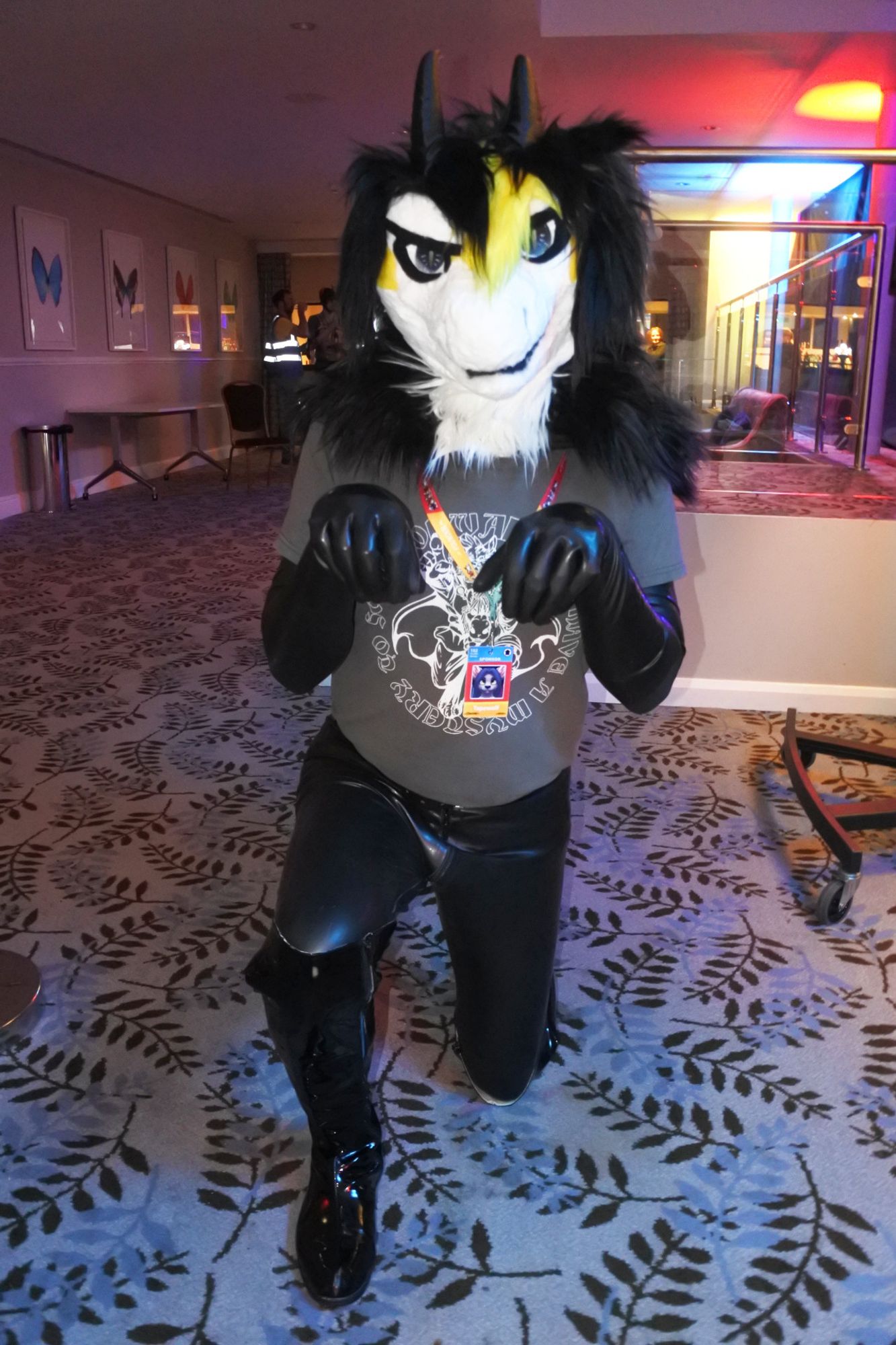 Fursuit photo of a black white and yellow dragon at Pawsome.  He is wearing latex opera gloves, latex leggings and pvc boots.
He's been asked to do a "puppy pose" by an onlooker

Photo by Mutley