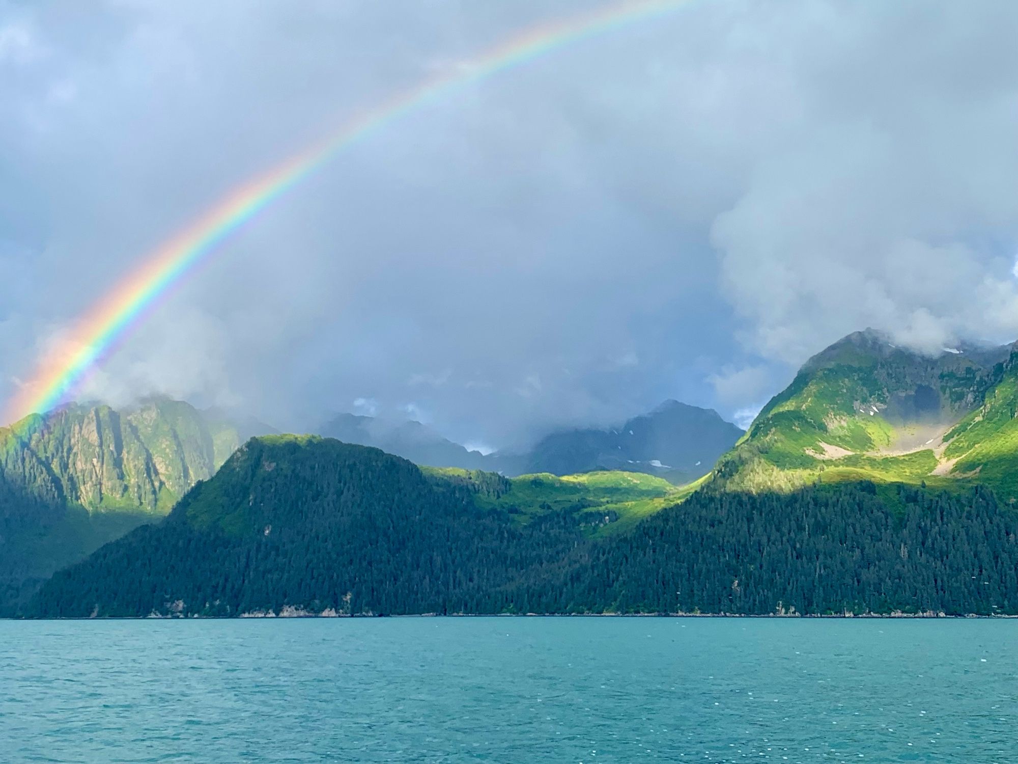 A rainbow over a vibrant green forested, mountainous landscape, with a teal blue sea.