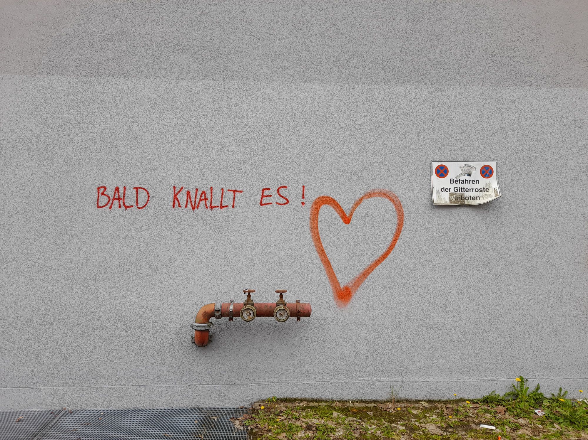 Graffiti in rot an weißer Hauswand: "Bald knallt es!" Rechts daneben ein großes rotes Herz.