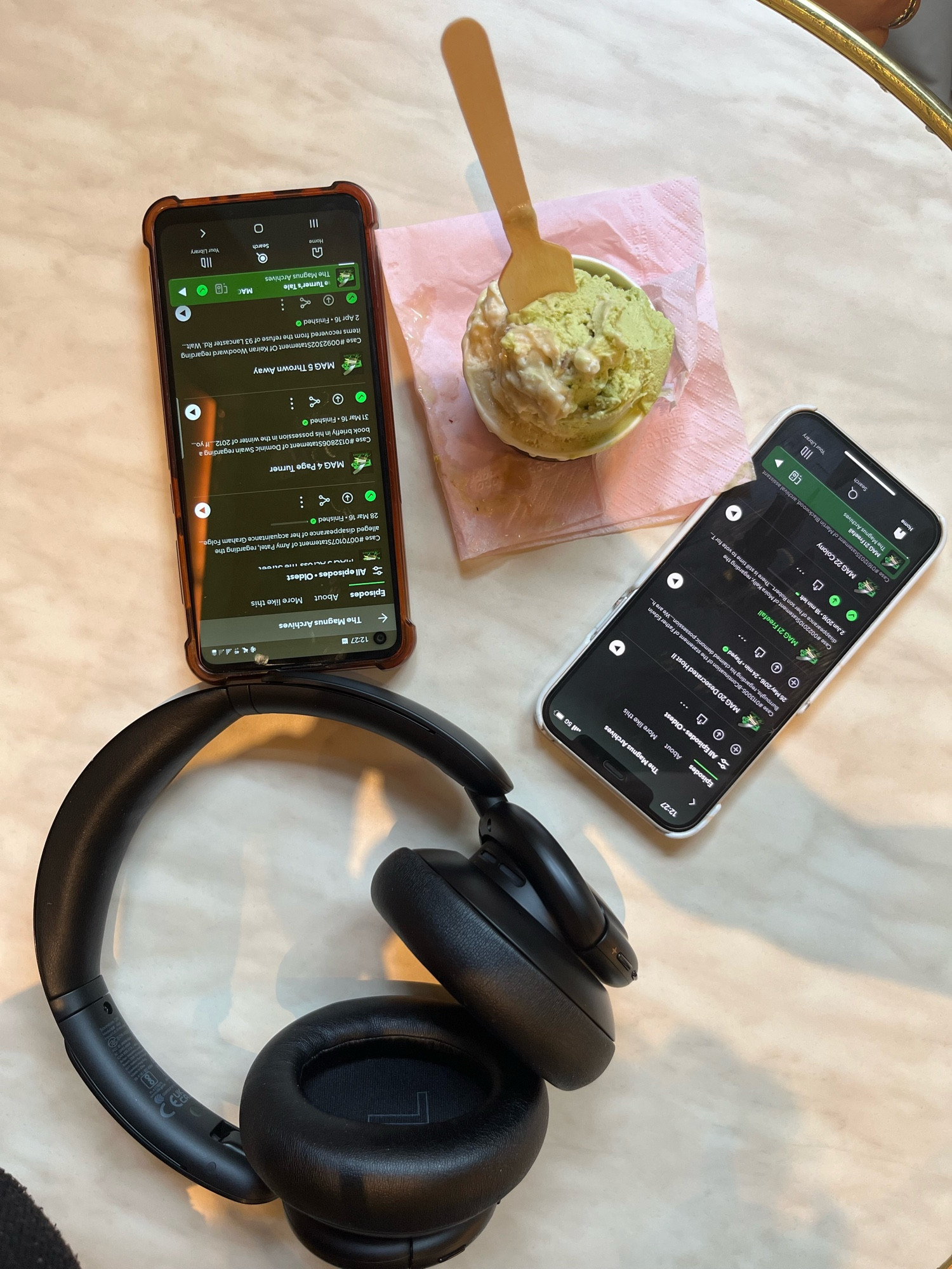 A pair of headphones and an ice cream cup are on a marble-like tabletop alongside two phones displaying The Magnus Archives episodes list on Spotify.