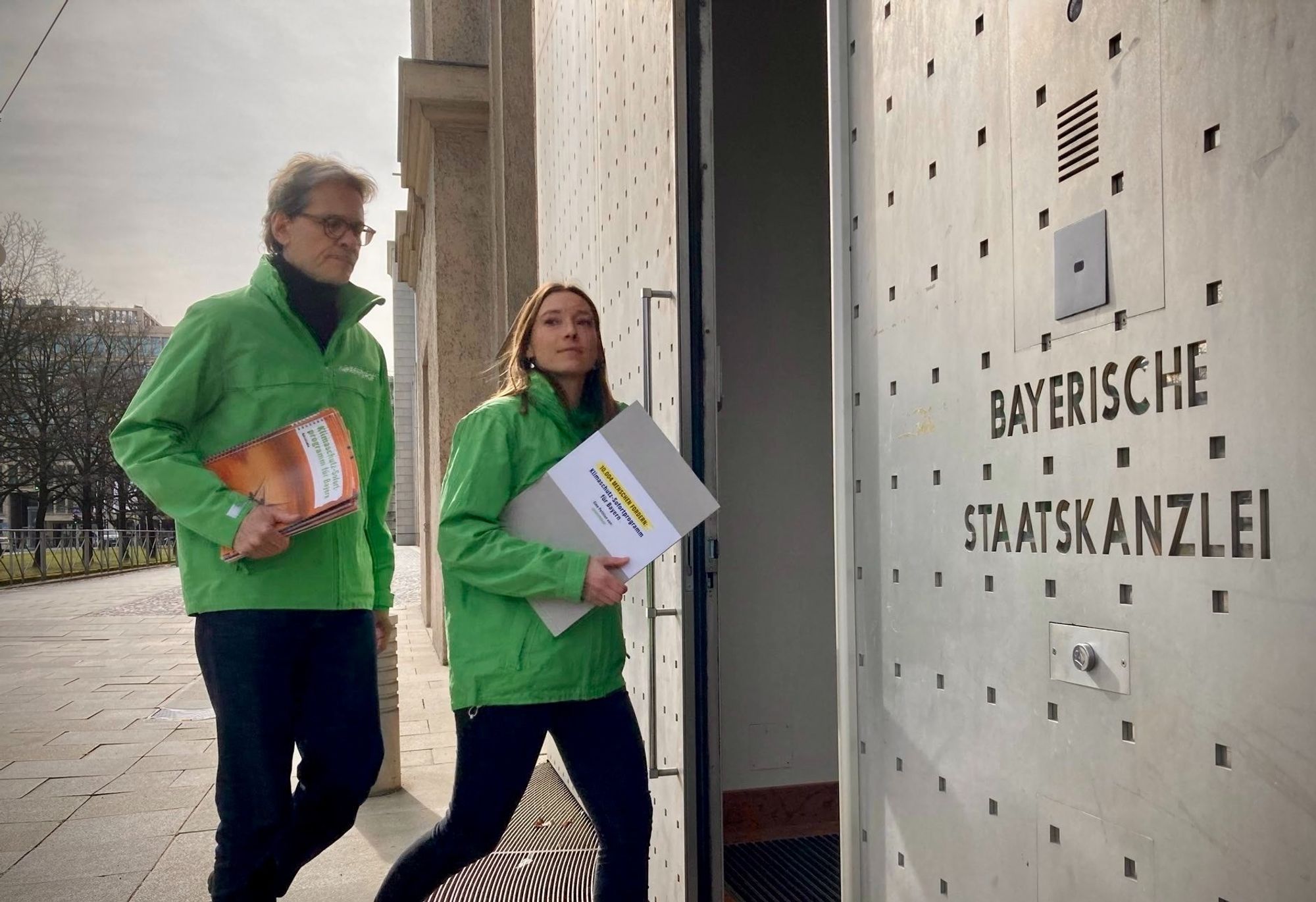 Greenpeace Aktivist:innen mit grünen Jacken bringen das Klimaschutz Sofortprogramm für Bayern und die Unterschriften in die Bayerische Staatskanzlei. Mann und Frau mit grünen Jacken und Dokumenten unter dem Arm betreten die Staatskanzlei.