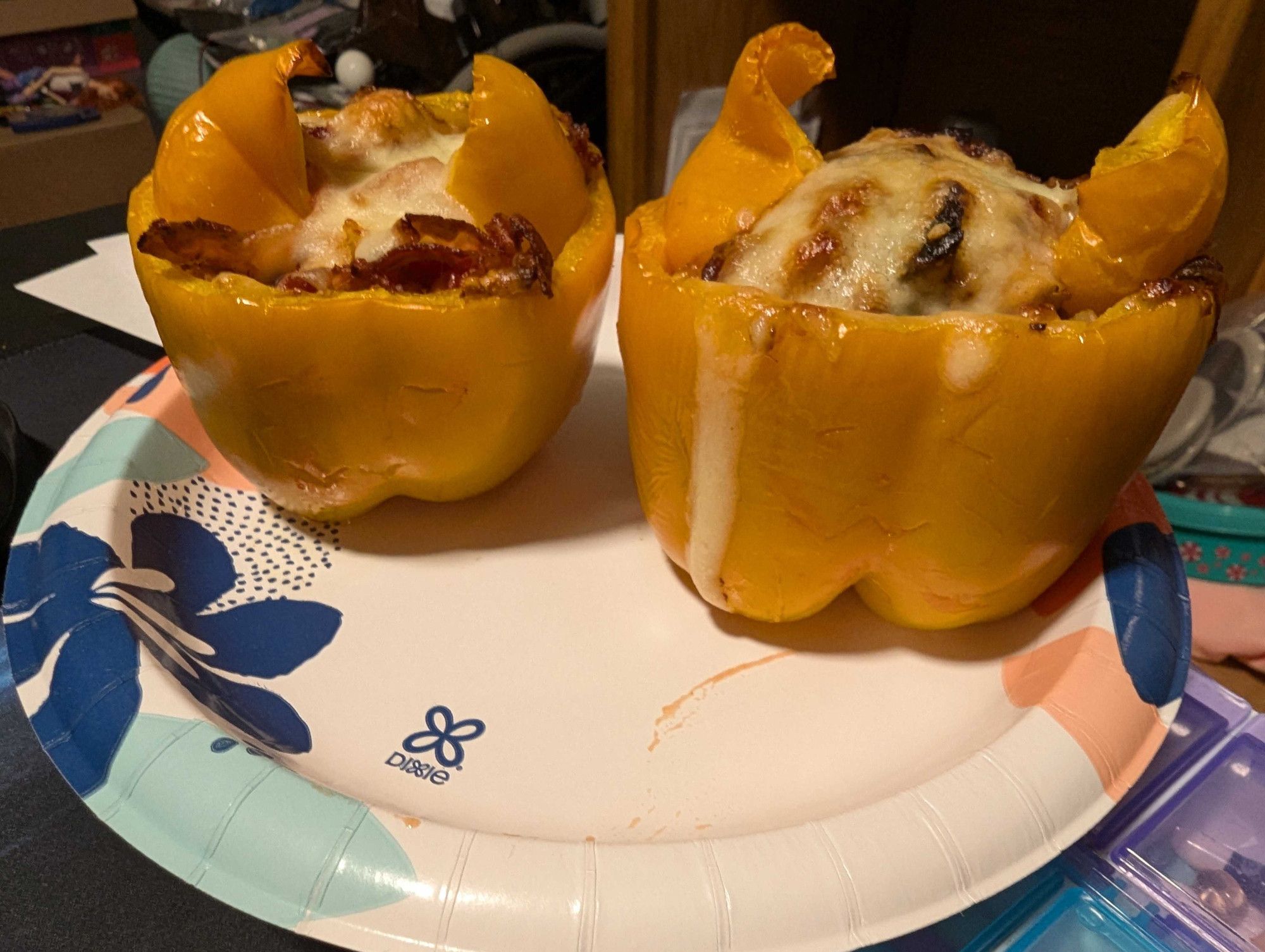 Two yellow bell peppers sitting on a plate.

The top of it has been slice off and stuffed with a mix of chicken, cheese, and bacon.

It's not super visible, but it has a cute cat face carved into it.

It also has cat ears on top of it.

Can see a multitude of things in the background. Mostly, boxes (with a couple barbies on it), my old wheelchair (with my white cane sitting on it), and our oak entertainment center.

Underneath the plate, can see my pill container and several of the pills in it.