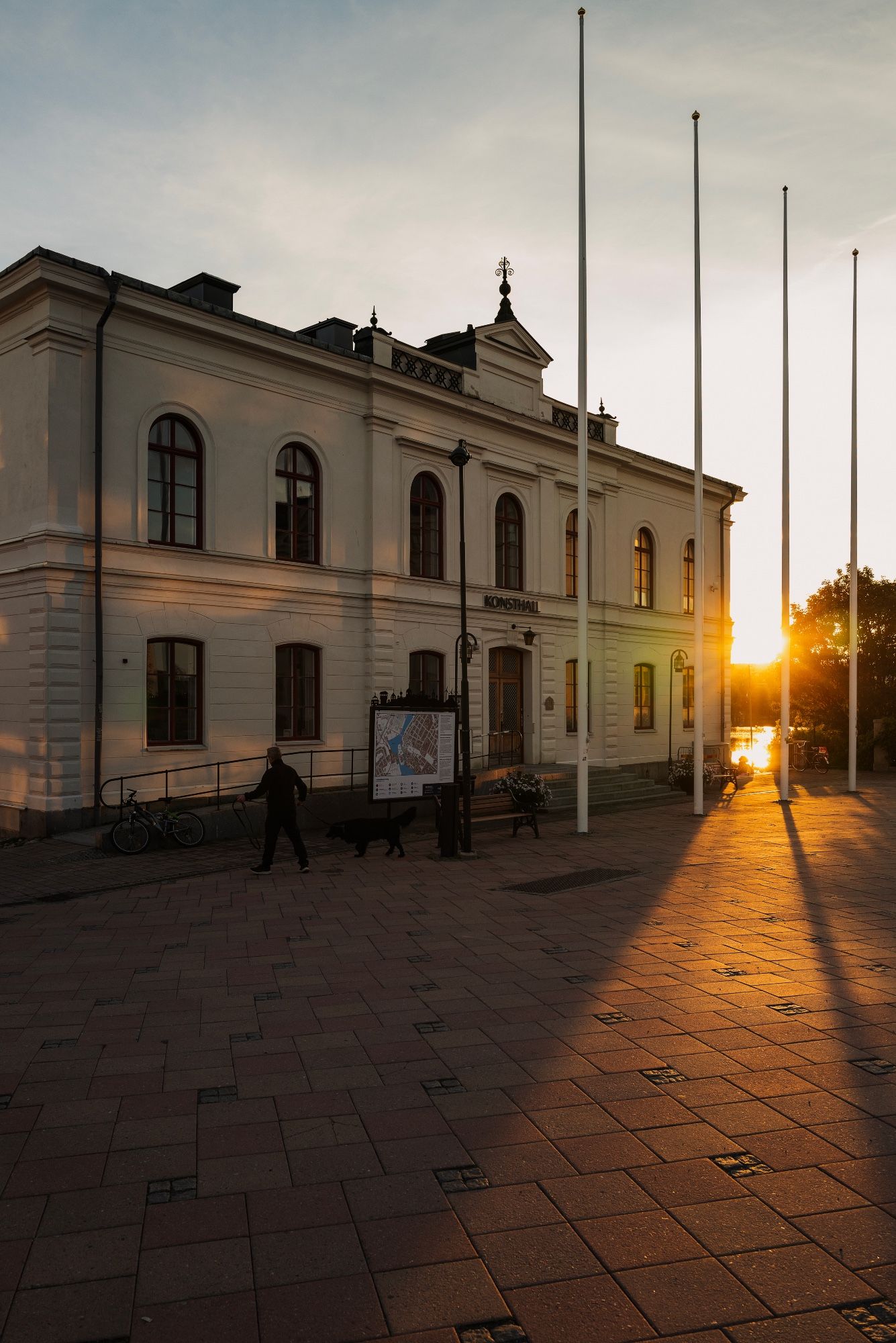 A museum building with a city square in front, some flag poles and a man walking his dog in front of the building. The sunset shining strong sunlight passed the corner of the building, the flag poles casting shadows.