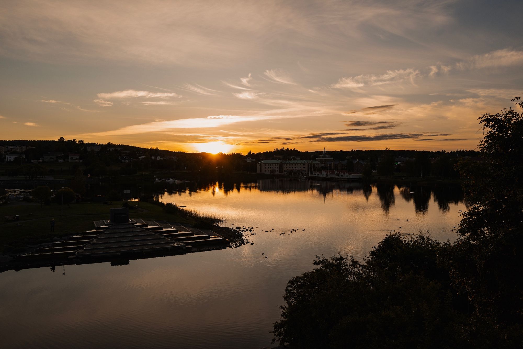 Sunset over a coastal town in Northern Sweden. Lightly cloudy skies, trees, buildings and the sunlight reflecting in the water. Ducks swimming in the middle of the frame.