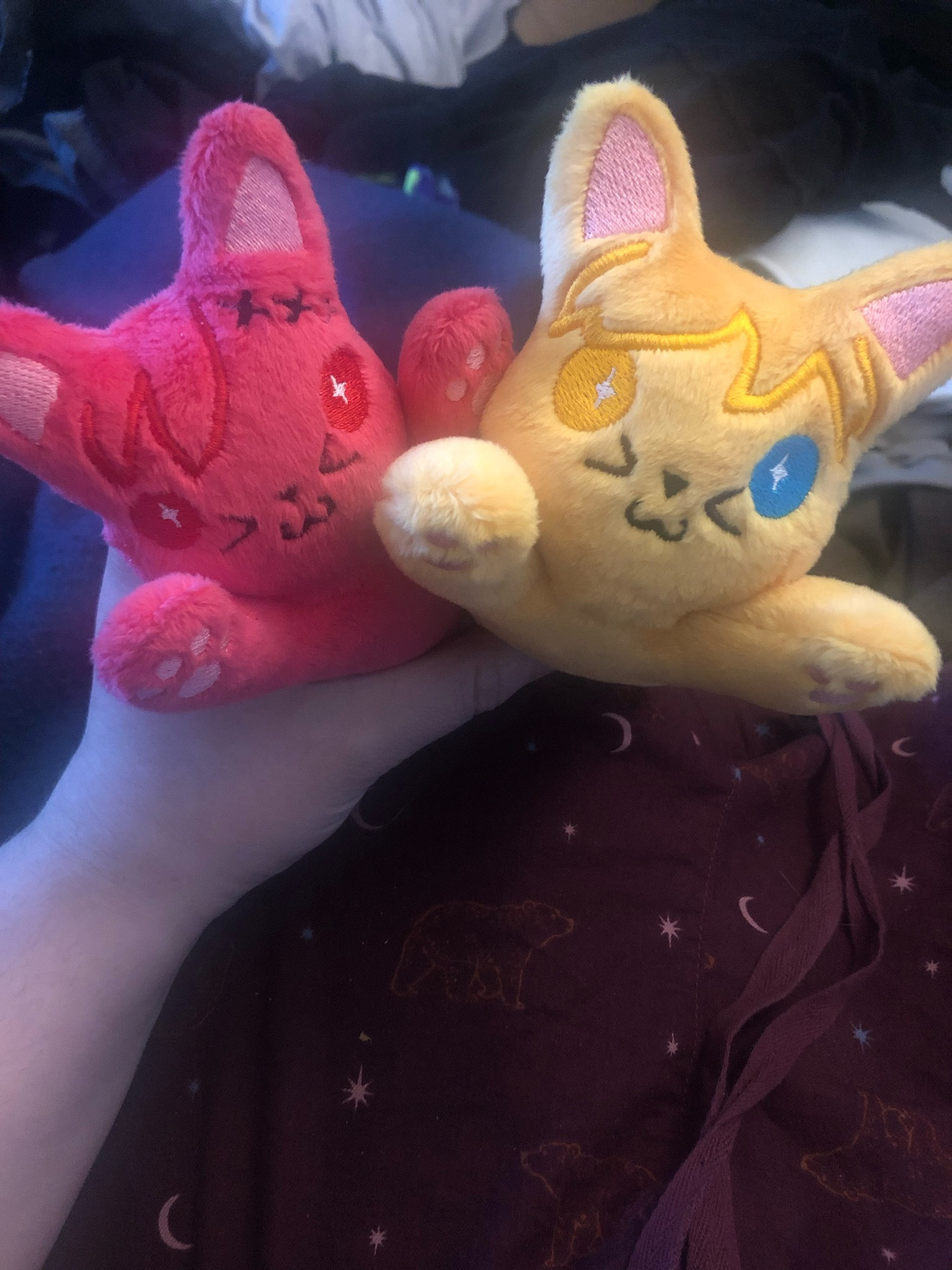 Two small cat plushies held in one hand. One is made with red fabric and red thread and has three black x’s on its embroidered hair, clearly made to resemble G’raha Tia from Final Fantasy 14. The other is made with yellow fabric and yellow thread, but one eye is a bright blue.