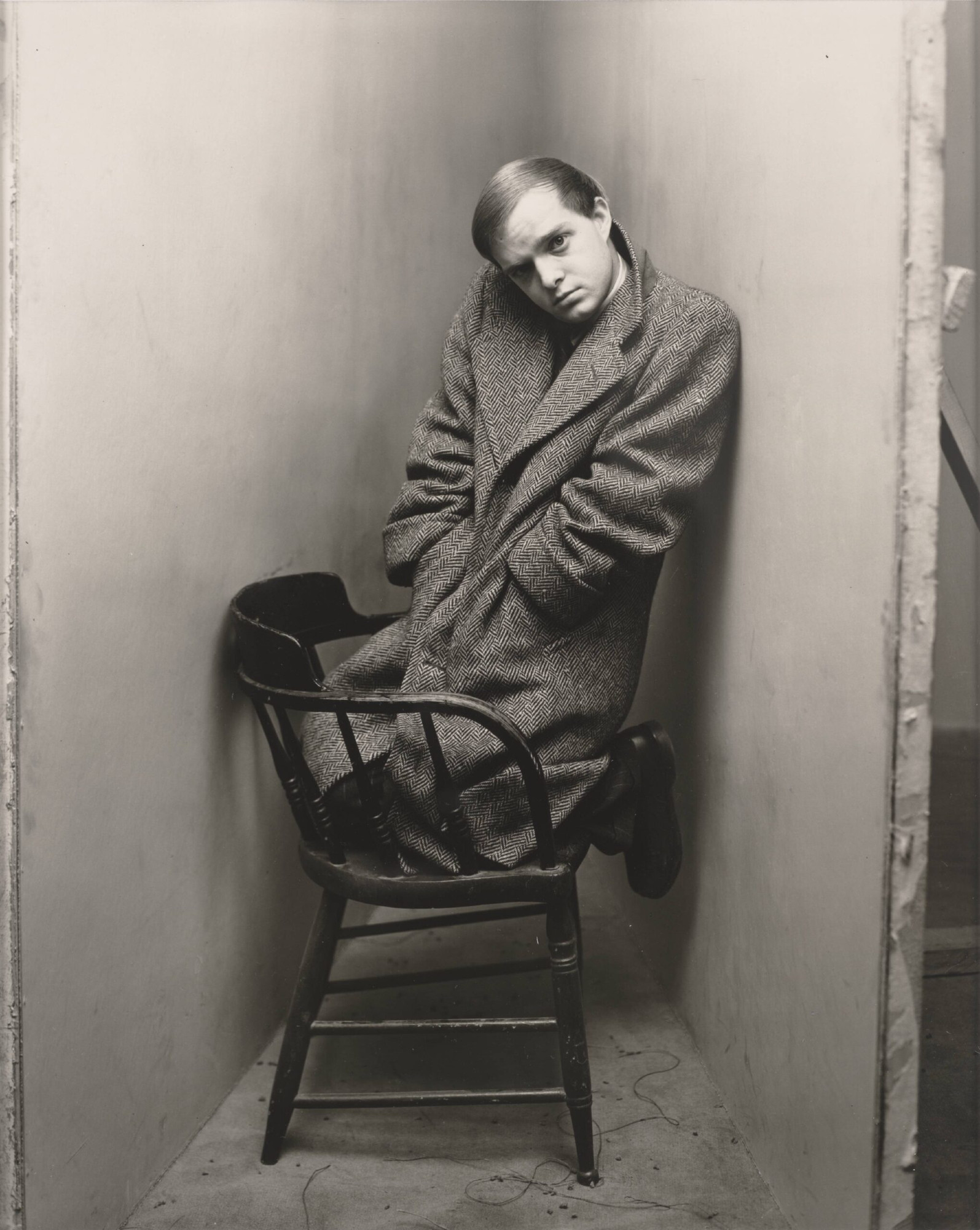 A photograph of writer Truman Capote. He's been placed in a tight, constructed corner, his knees resting on a wooden chair, enveloped in a heavy coat, his head cocked as he stares out at us