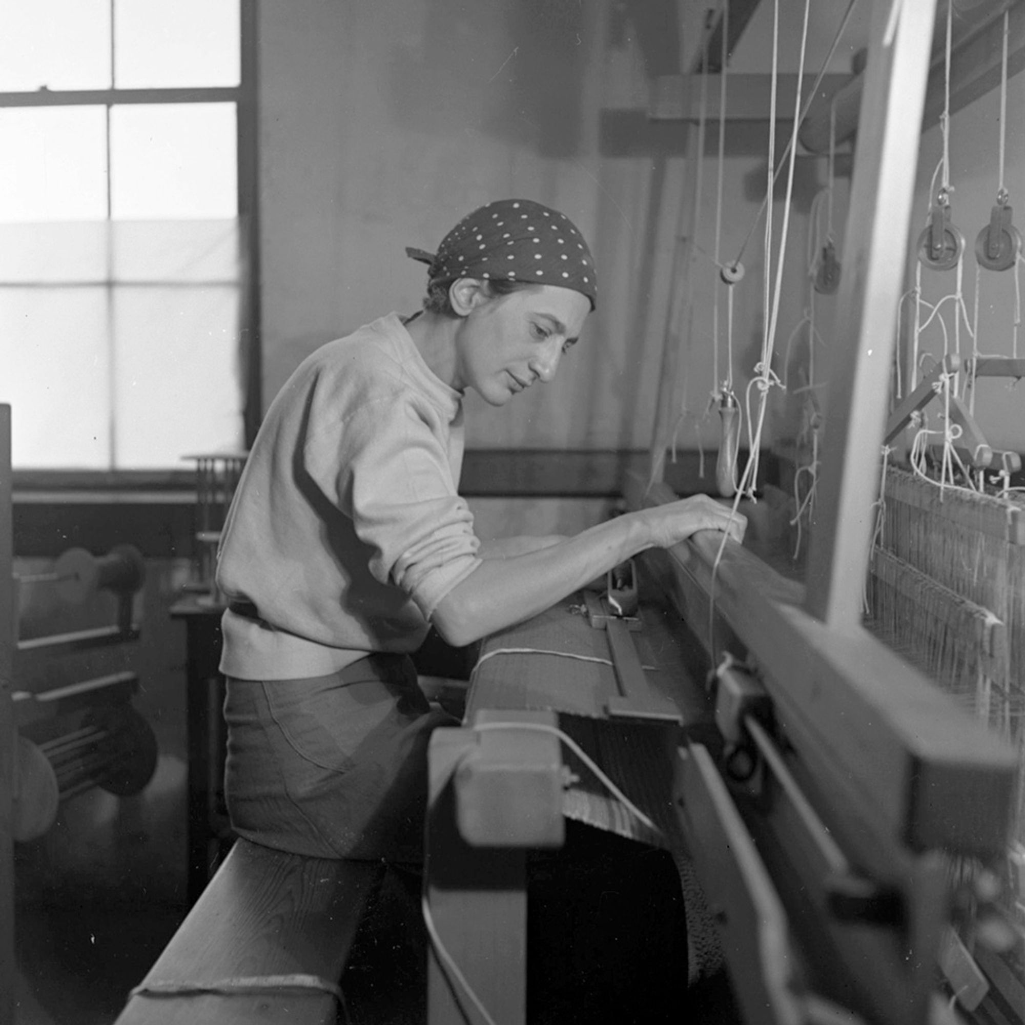 A black-and-white photo of a woman at a weaving loom, seen from the side, a bandana or kerchief pulled tight around her head