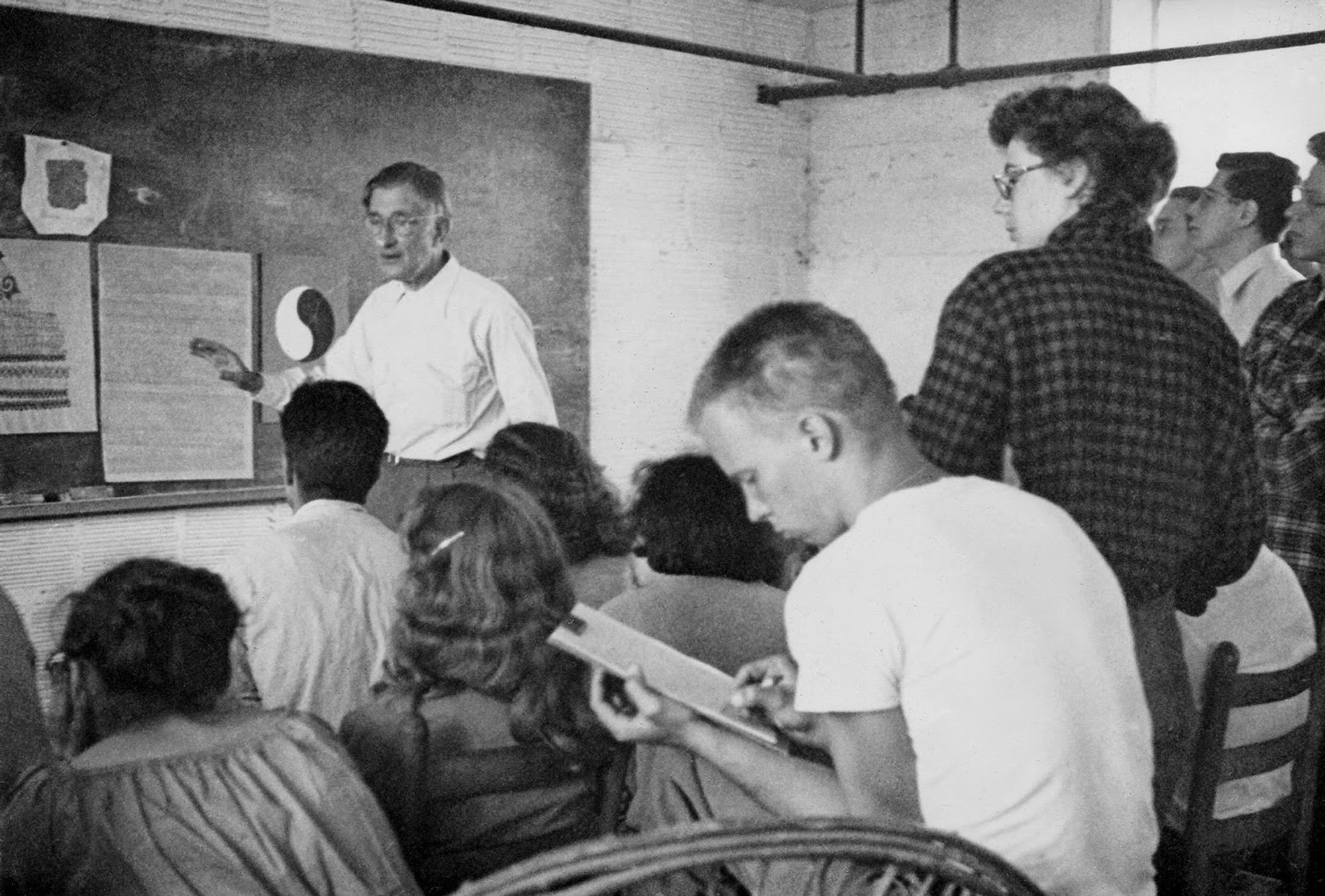 A classroom setting. An older man stands before a group of students, gesticulating, apparently lecturing to them. In the foreground a young, sandy-haired man in a white t-shirt looks to take notes