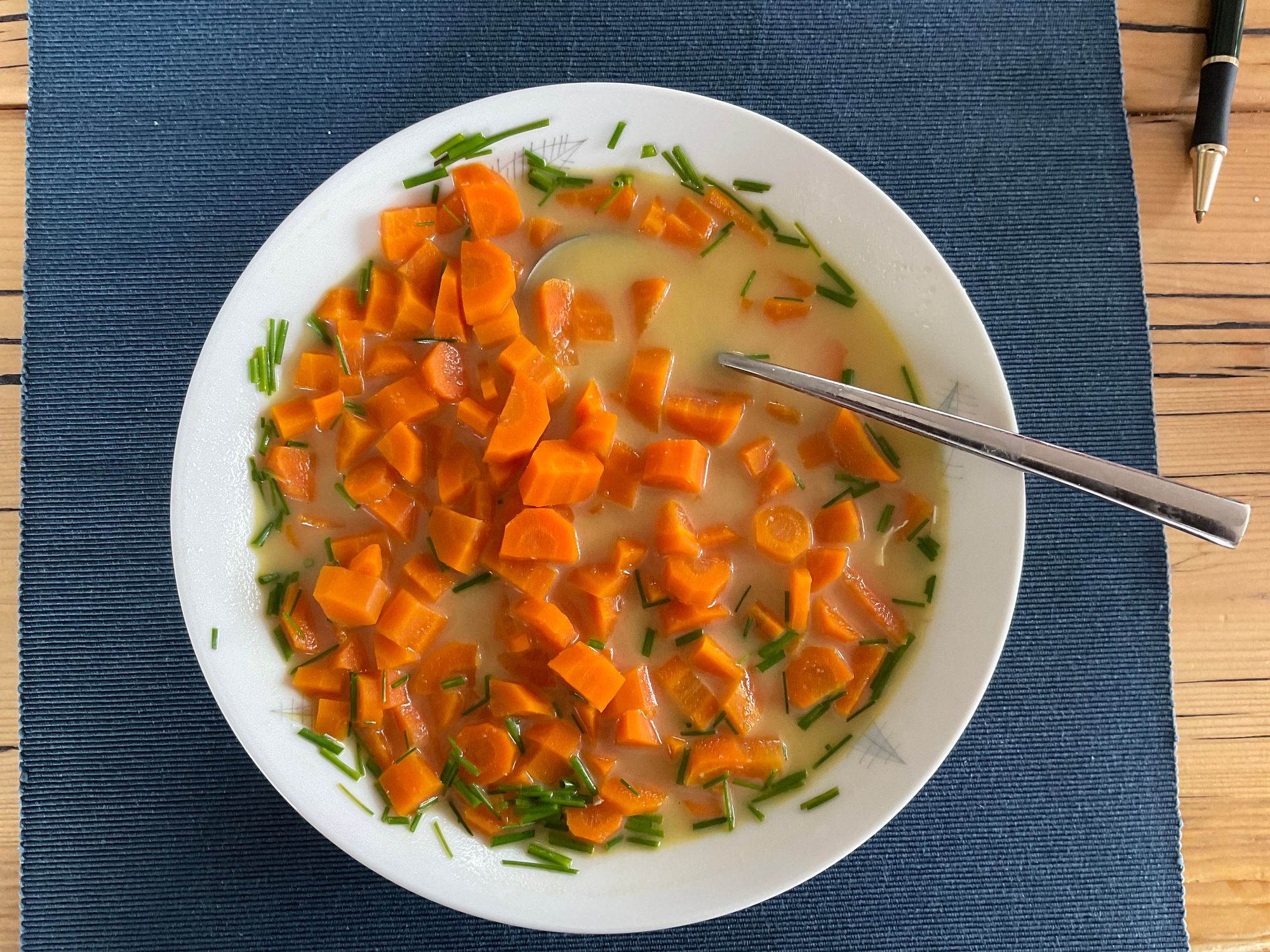 Eine Schüssel Suppe mit gewürfelten Karotten und gehacktem Schnittlauch in einer cremigen Brühe, serviert auf einem blauen Tischset. Ein silberner Löffel ruht in der Schüssel.