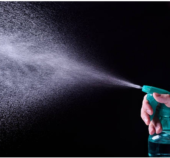 White person’s hand spraying a stream of water from a transparent green spray bottle.