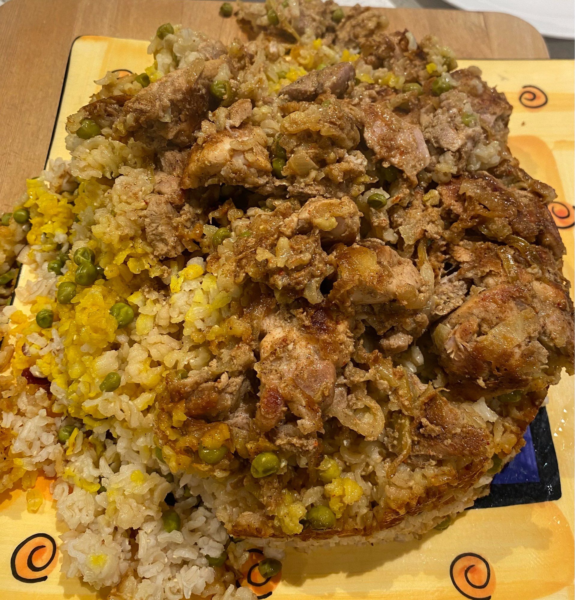 Brown rice chicken biryani on a 90s yellow hand-painted square serving dish. At left some of the dish has tumbled off from the main form of the rice. There are some peas and saffron-stained rice. Pieces of marinated chicken on sitting on the top.