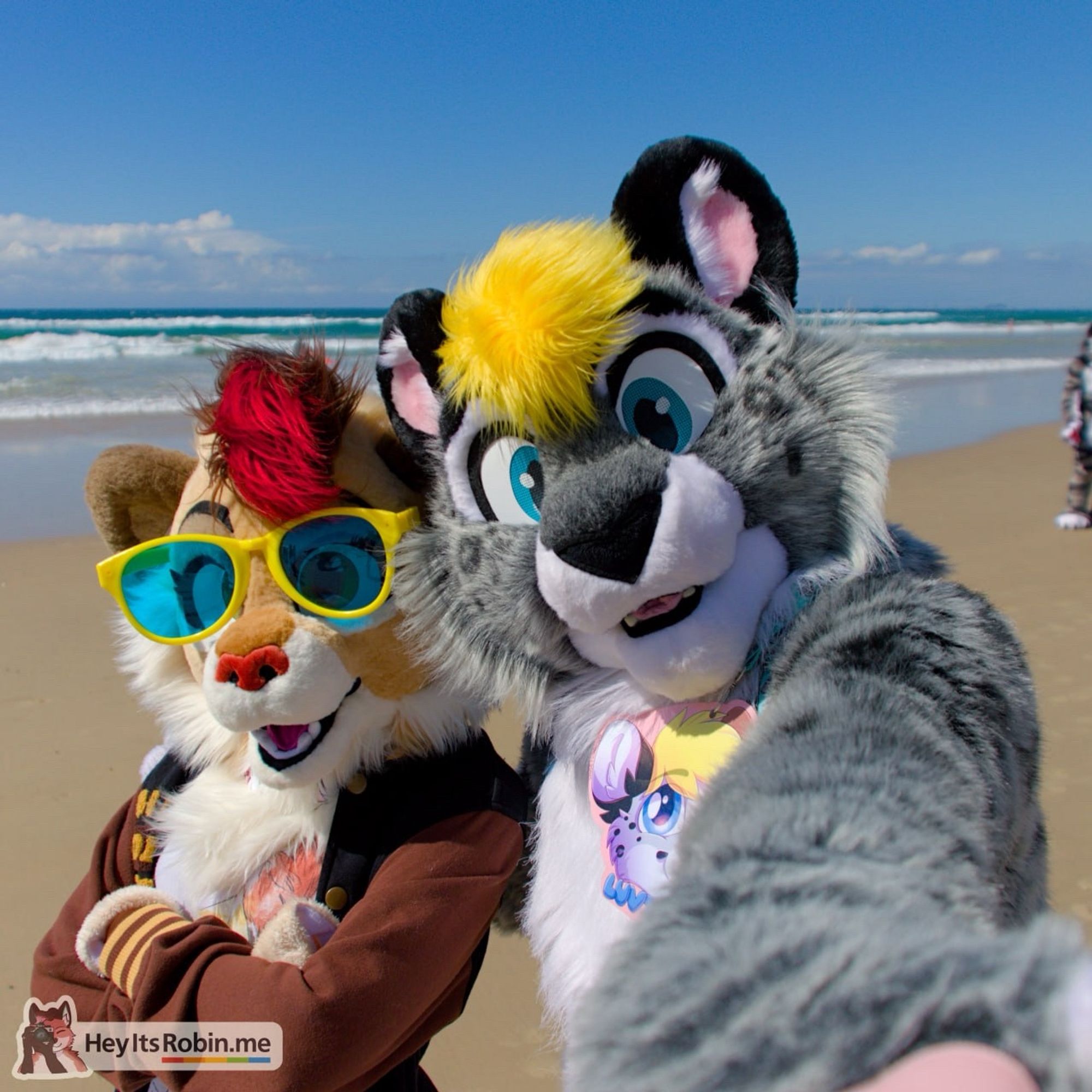 Lovetail, a snow leopard fursuit, and Sneaky, a lion fursuit, stand on a beach posing for a photo. Lovetail’s paw is outstretched towards the camera, giving the illusion of the photo being a selfie.