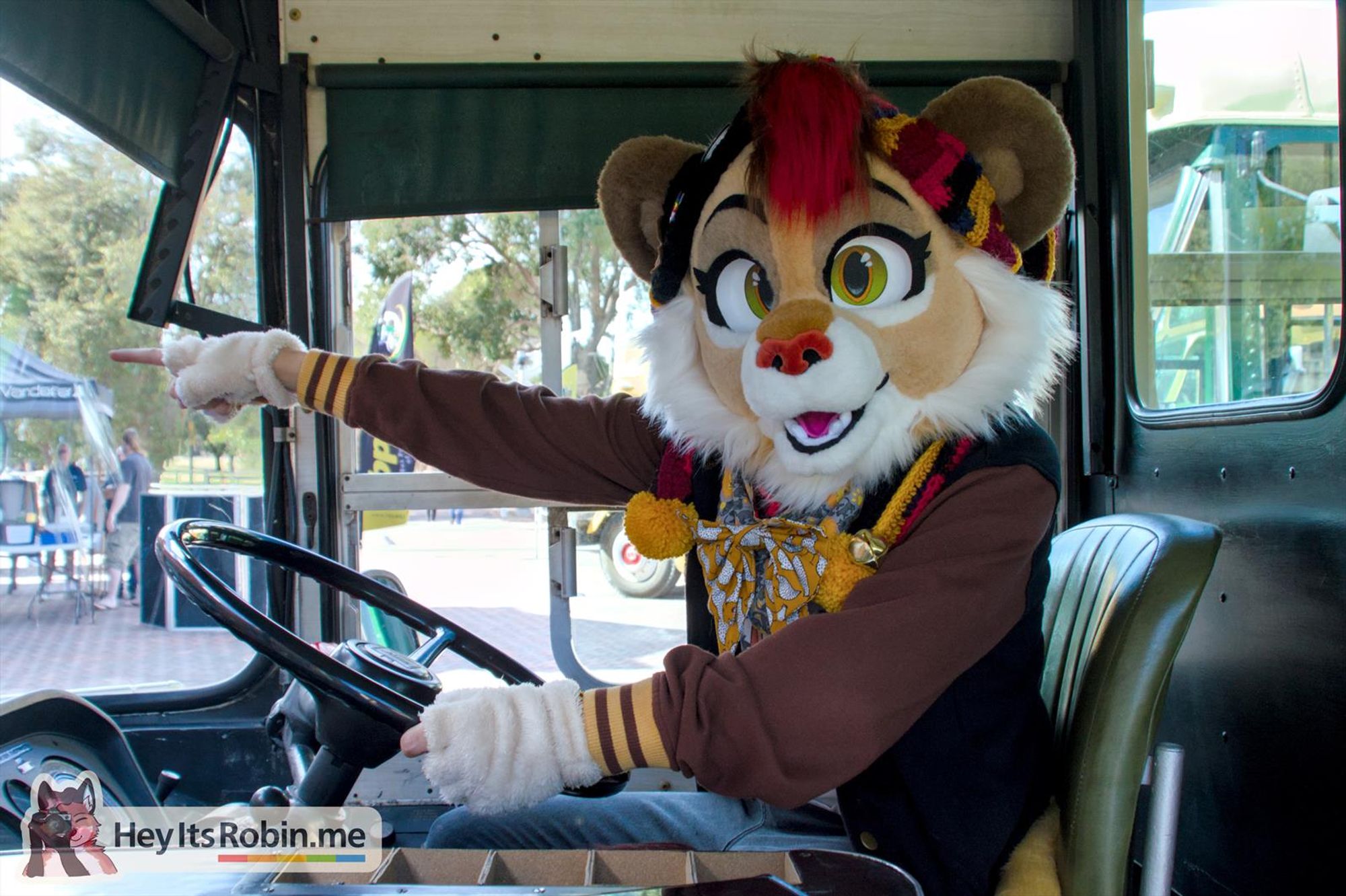 Sneaky, a lion partial fursuit wearing a varsity sweater, sits in the driver's seat of a vintage bus. She looks towards the viewer in the doorway, pointing forwards through the windscreen.