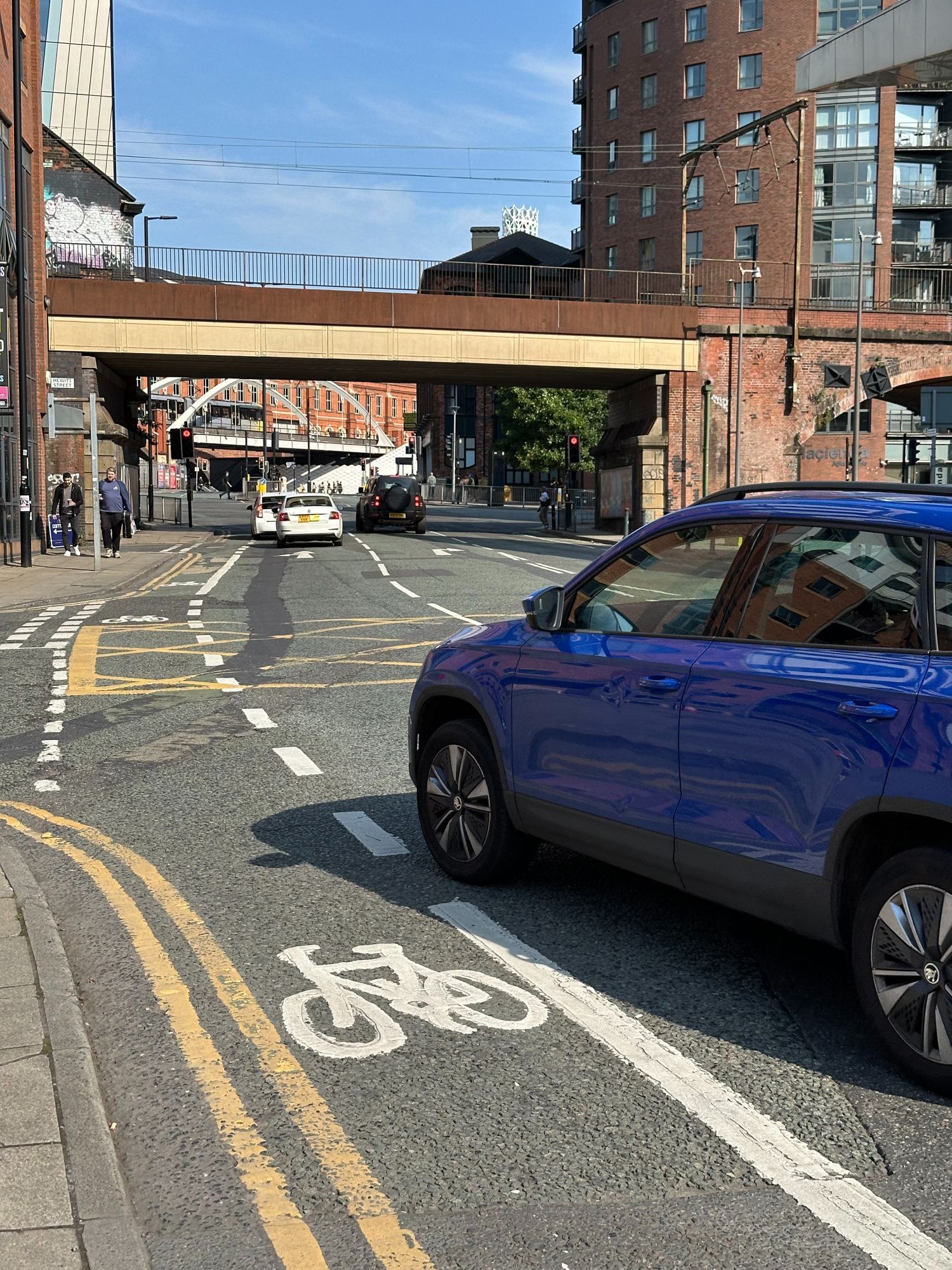 A painted bike lane in central Manchester with a car passing on the right, leaving very little space between the car and the kerb.