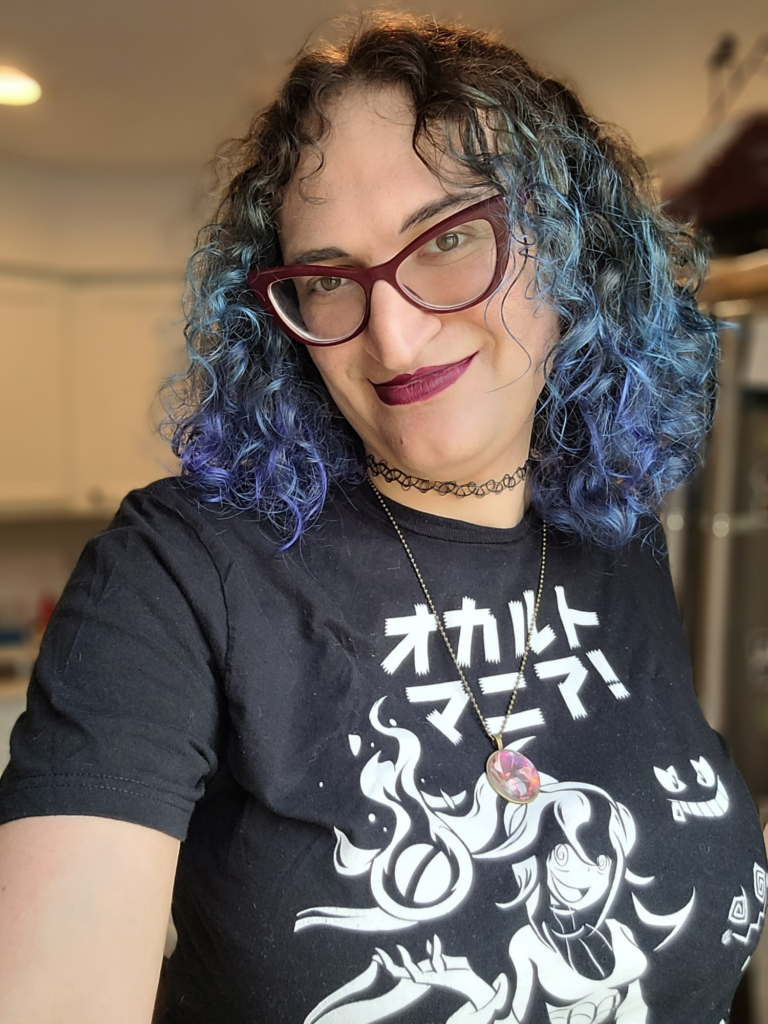 Becca, with long curly blue and indigo hair. She is wearing a T-shirt with the Hex Maniac from Pokemon, that says "OCCULT MANIAC!" in Japanese