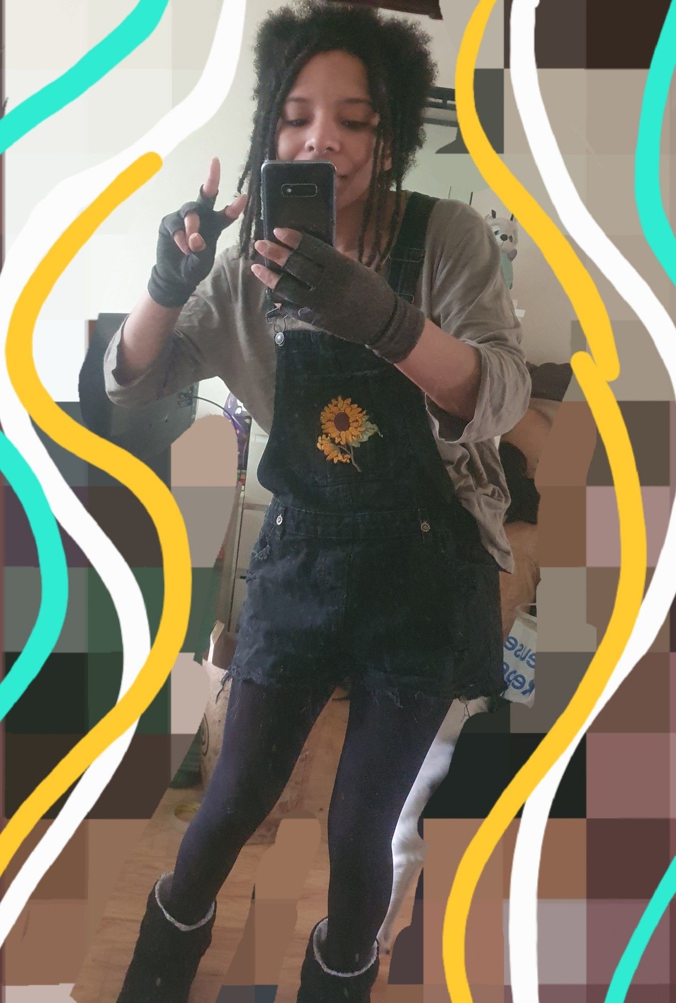 A mirror selfie I took of my full outfit showing off my dungarees I customised myself with sunflower themed patch and charms. I am also wearing  black tights, a lichen green shirt and doing another peace symbol with my hand.