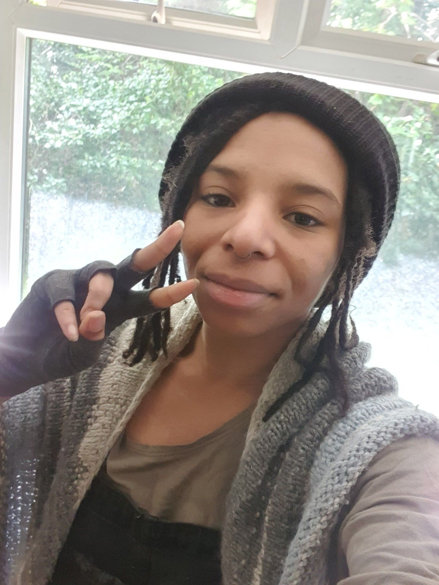 A selfie of me wearing a black beanie with a braided fringe and a blue/grey shawl draped over my shoulders doing a peace symbol with my hand.