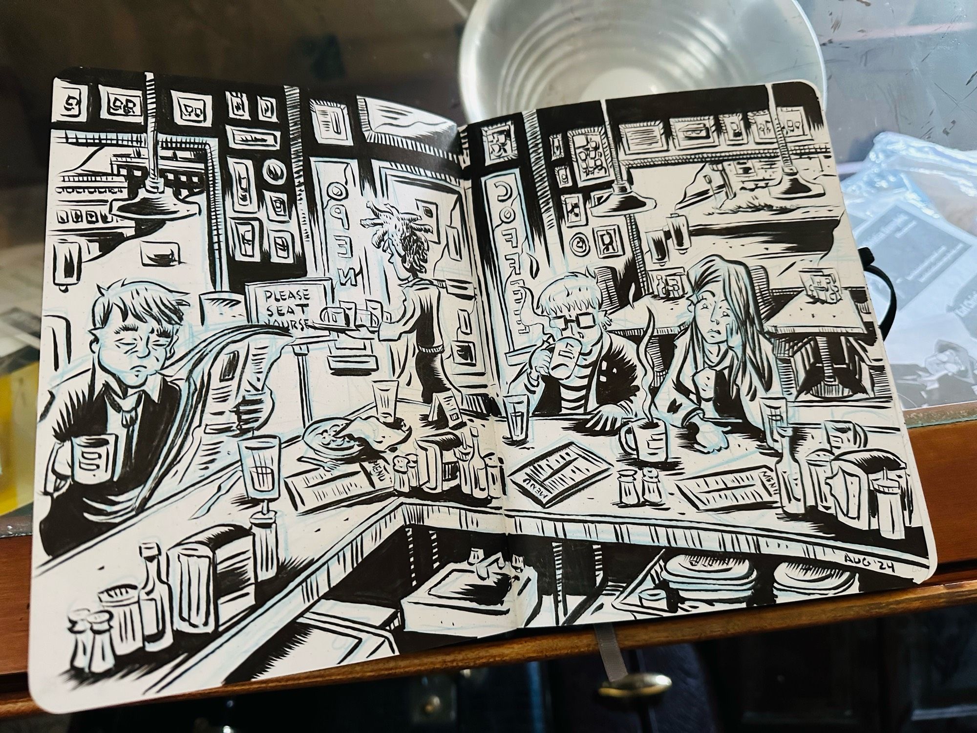 An open sketchbook on a drawing table with a two page spread B&W illustration of a pair of tired women sitting with coffee at a cafe counter.