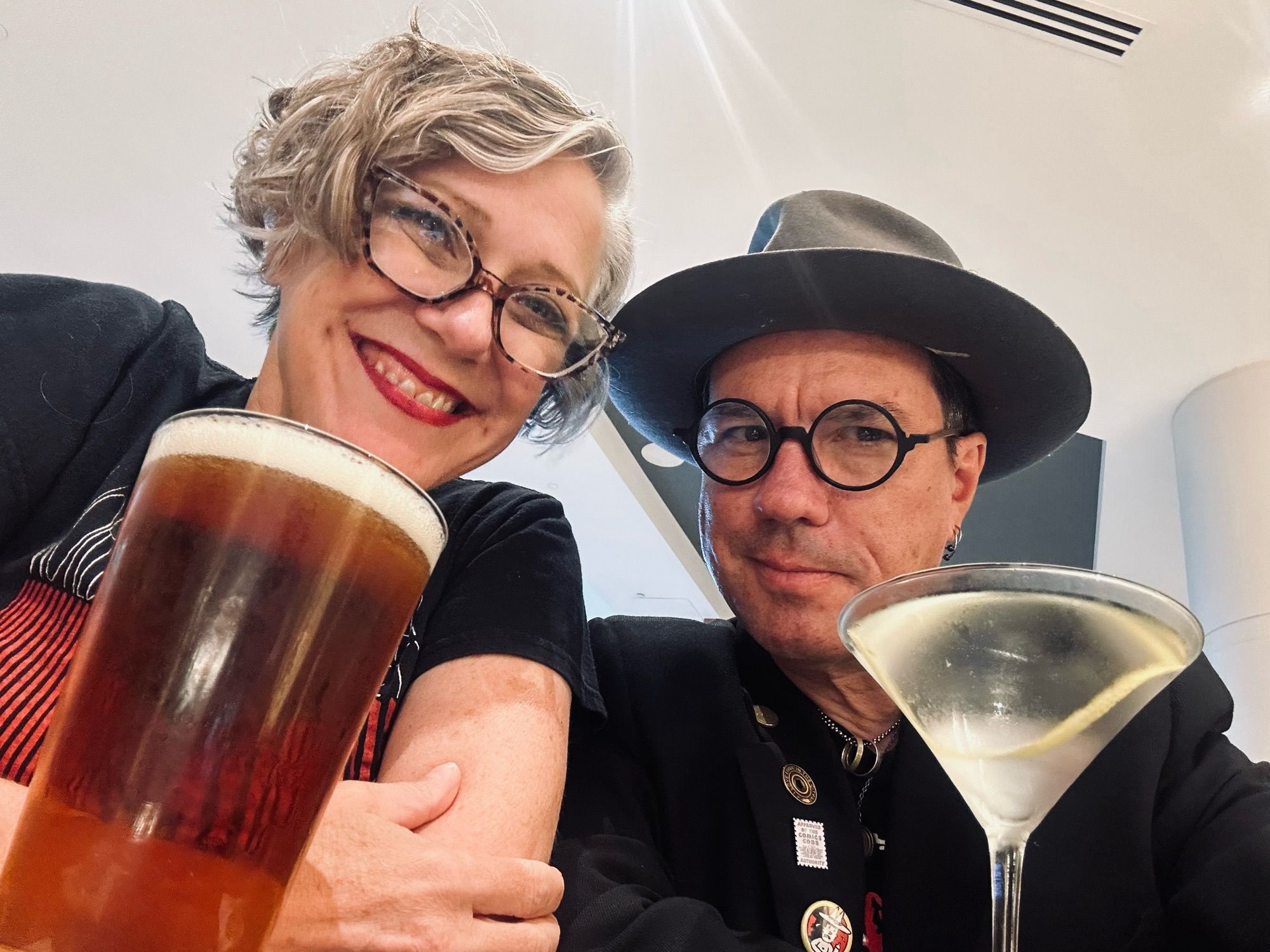 A man and woman, both wearing glasses, relax with a martini and beer.