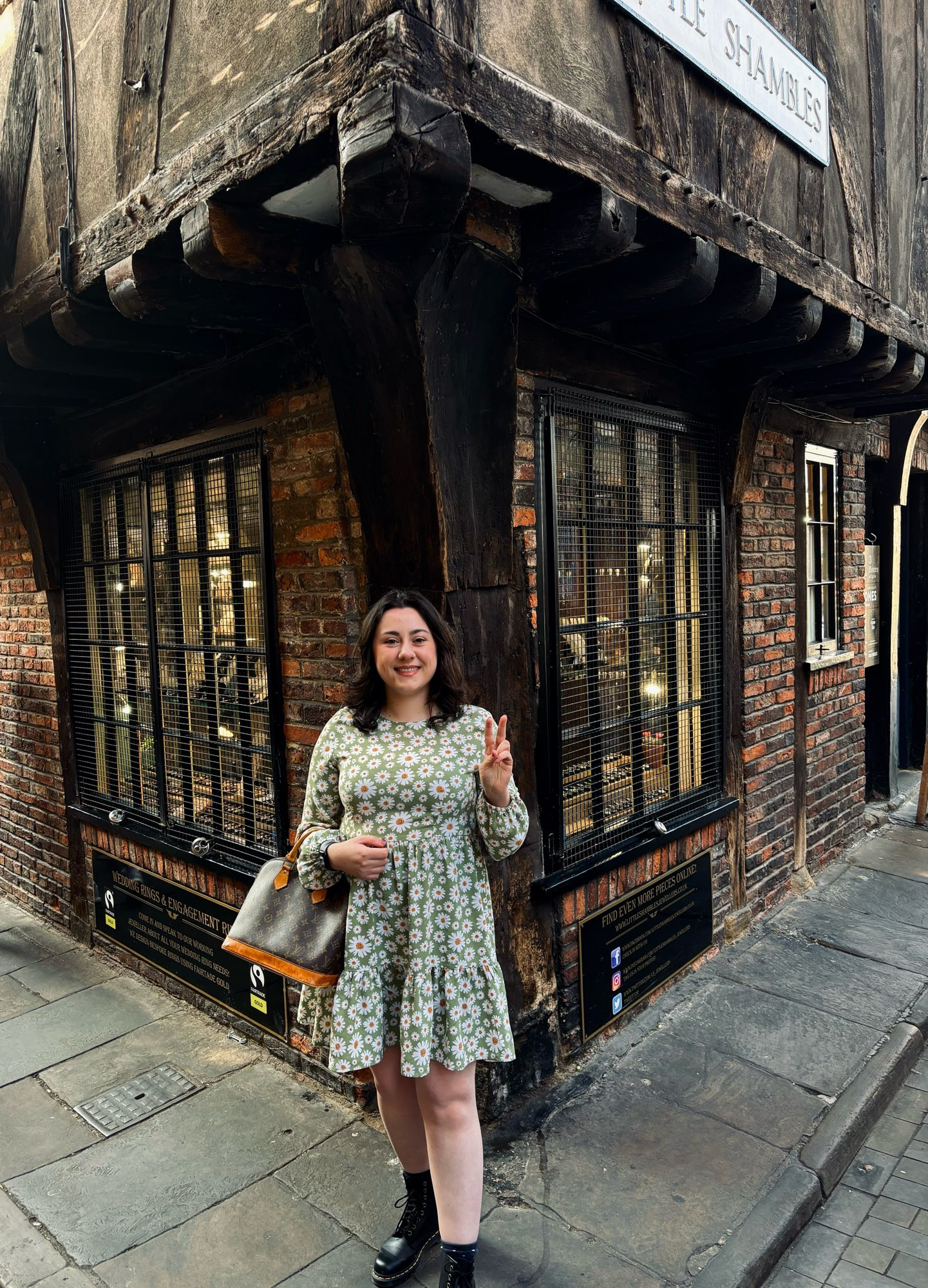 nosku stood under the shambles street sign doing the peace sign. she is wearing a green dress with flowers on it, a louis vuitton bag and doc marten boots. 