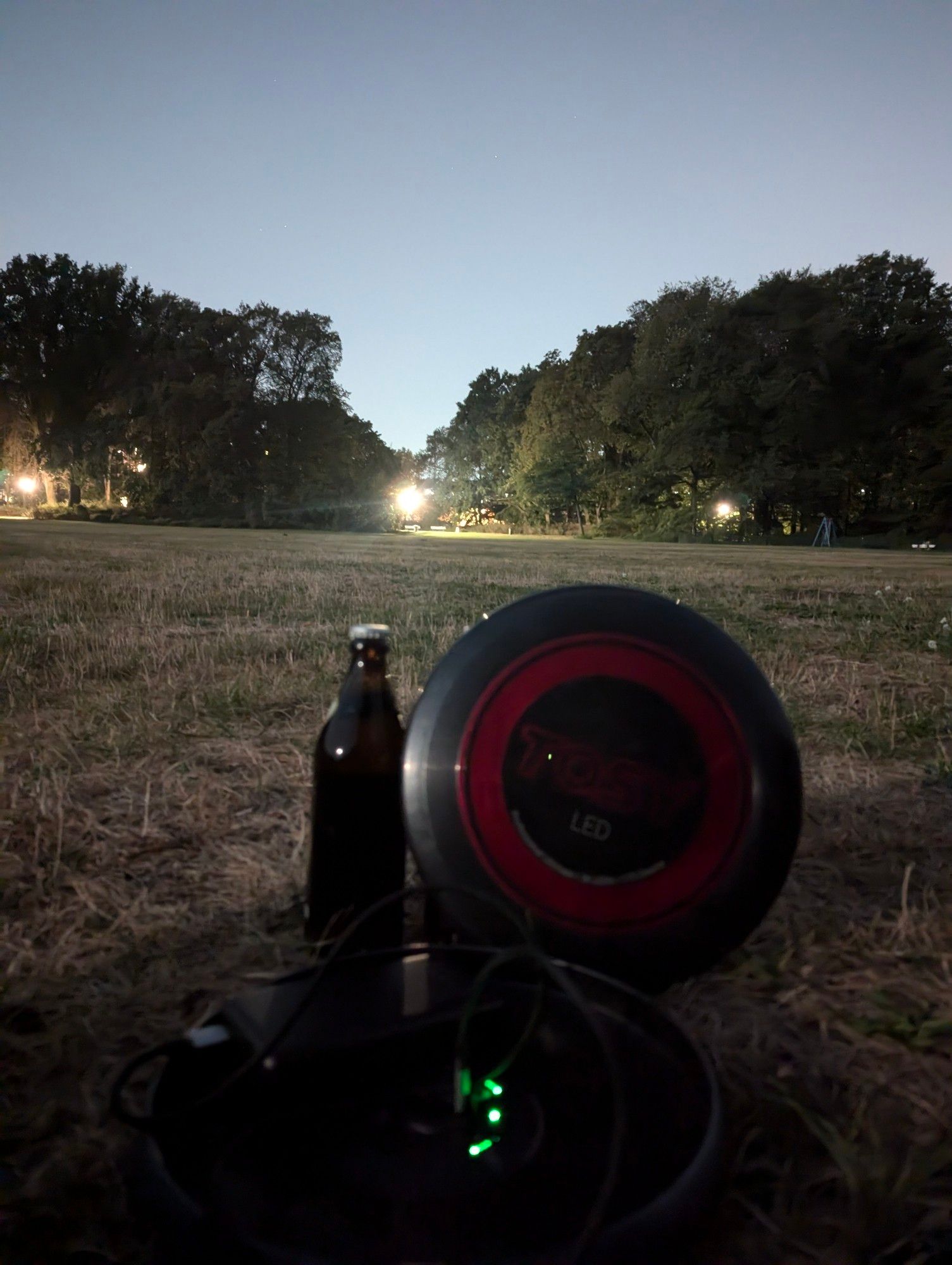 2 LED Frisbee, eine davon liegt und wird vom einer Powerbank aufgeladen, die andere steht, gegen eine Flasche gelehnt neben einer anderen Bierflasche. Im Hintergrund helle Lichter von der Nachtaufnahme.